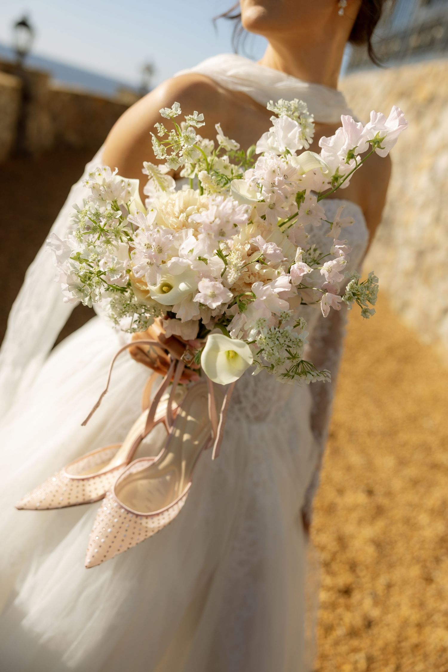 Bride in white tulle gown holding elegant bouquet of soft pink and white flowers with ribbon accents outdoors.