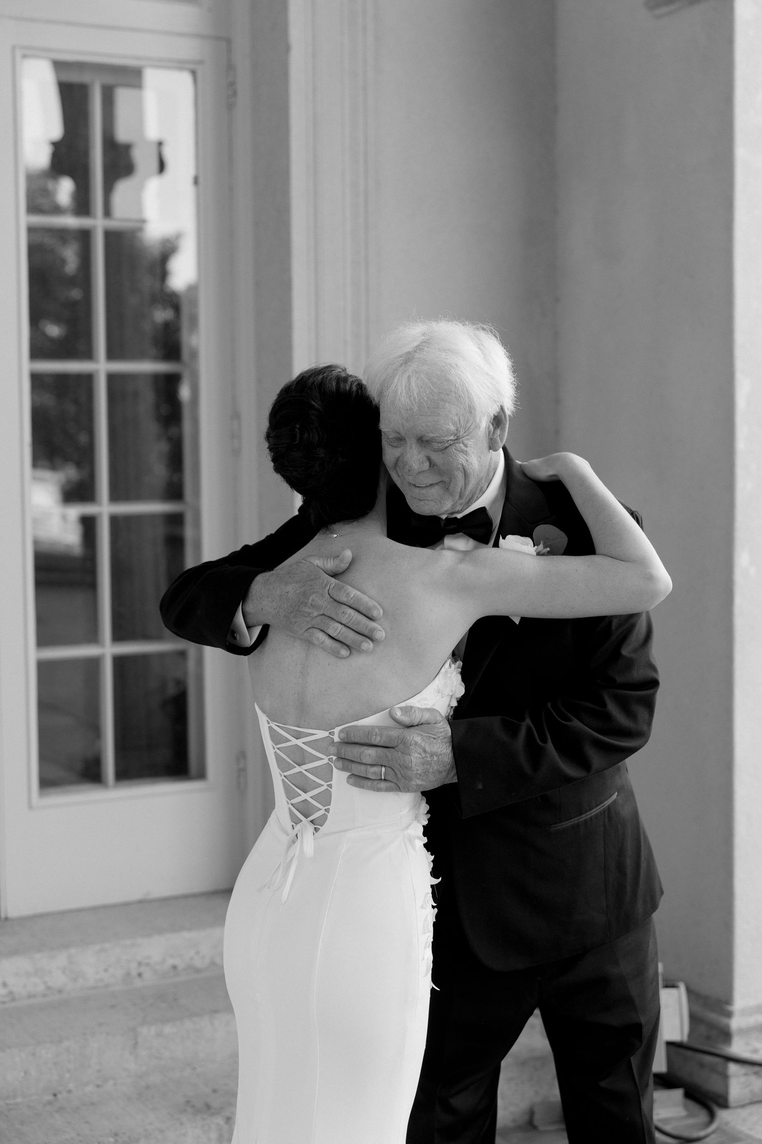 Emotional black and white photo of bride embracing elderly man in tuxedo at wedding venue.