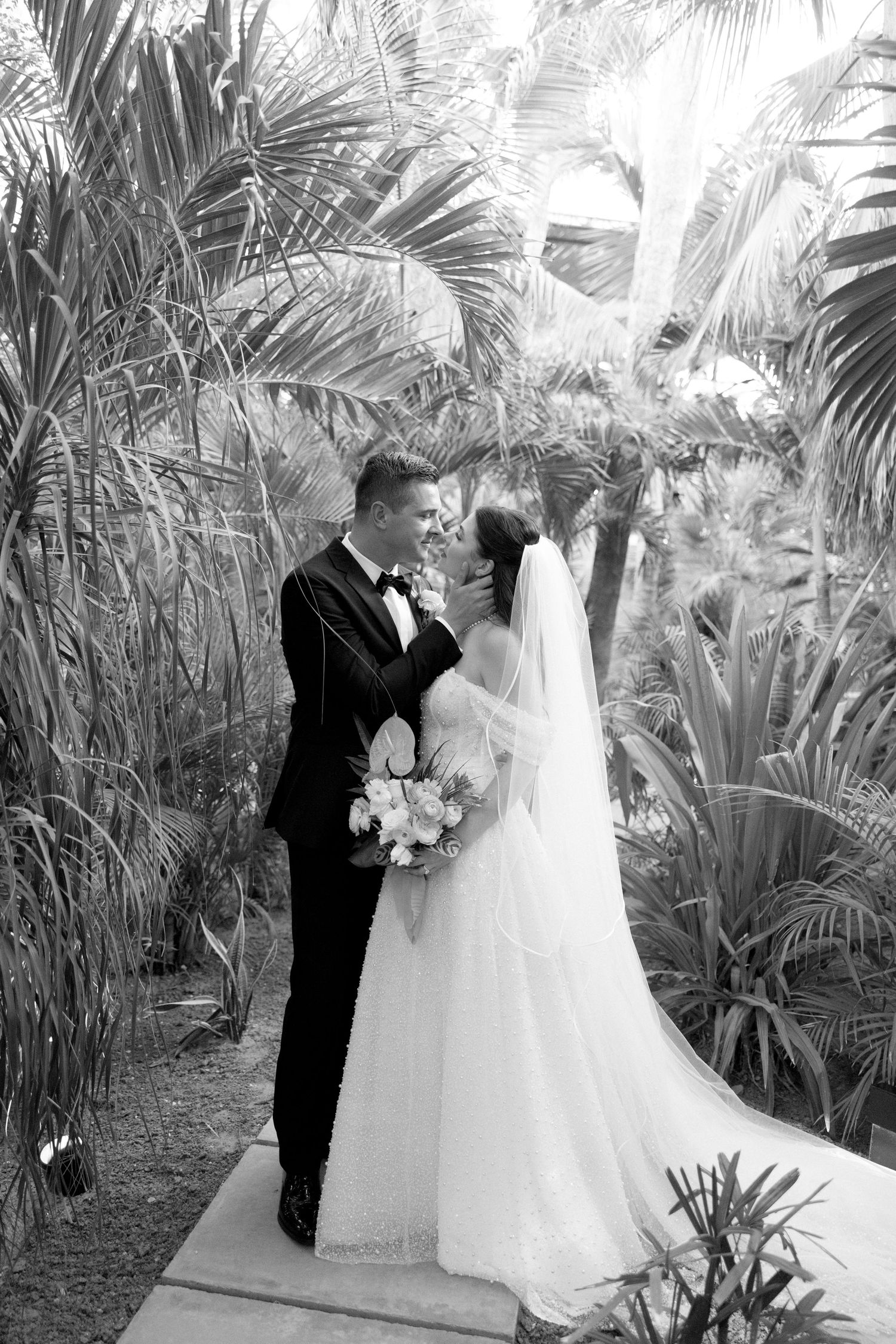 Black and white photo of bride and groom sharing an intimate moment surrounded by lush tropical palm foliage.
