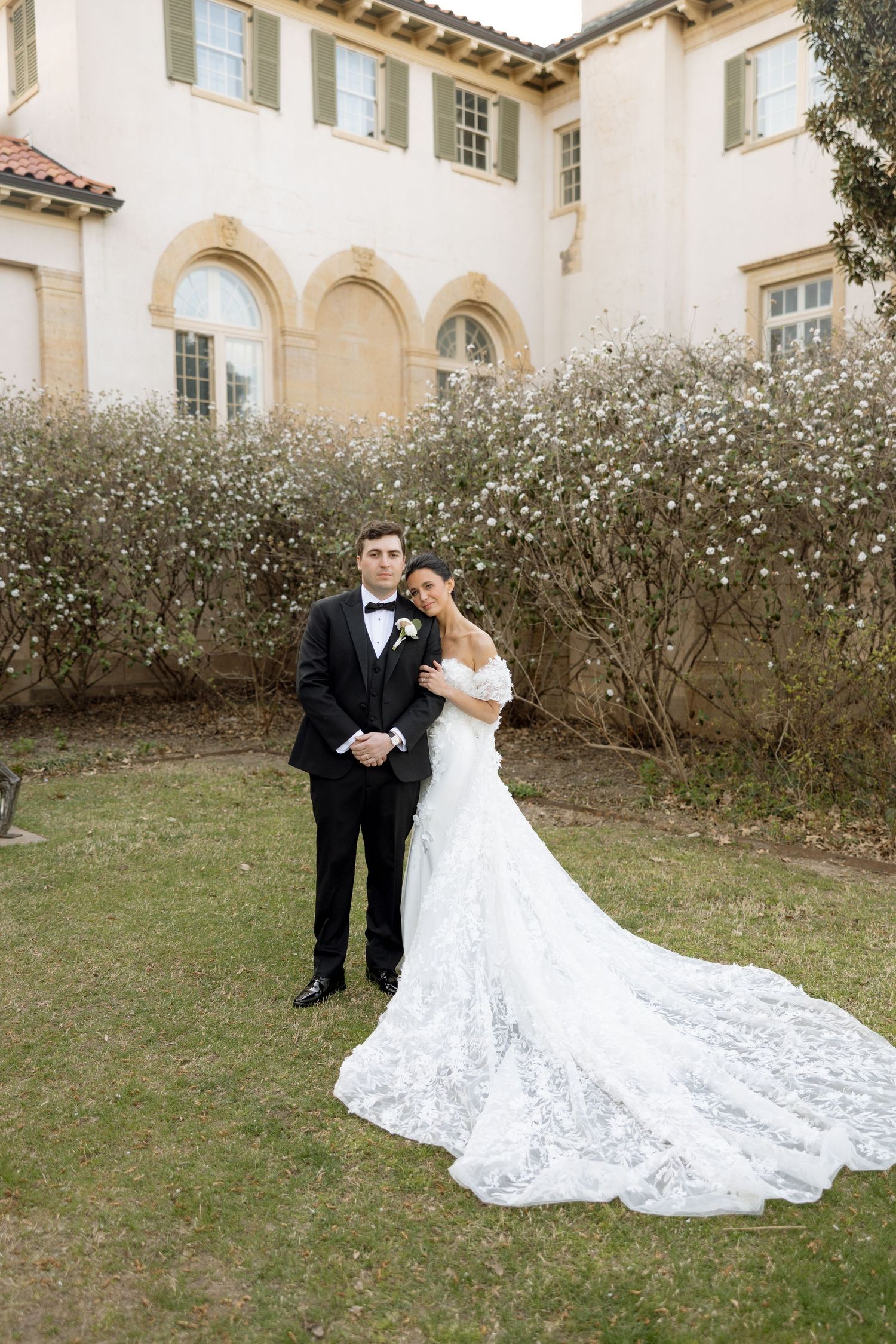 Bride and groom pose in front of an elegant estate, she wearing a lace mermaid gown with long train.