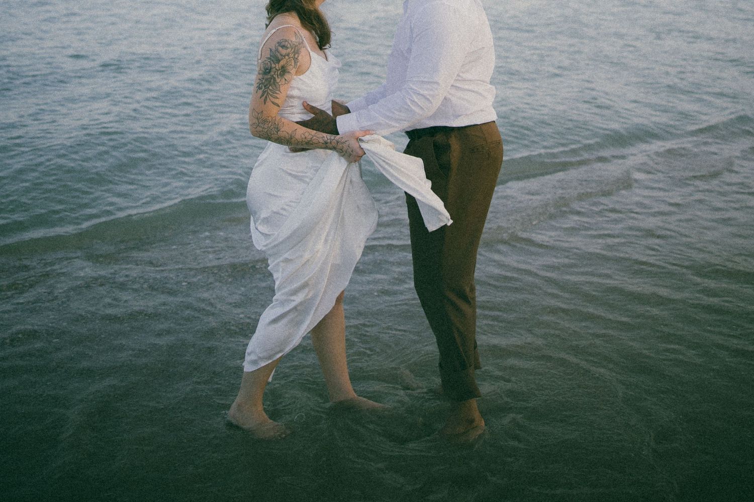 Couple playing in shallow ocean water, white dress flowing in the breeze.