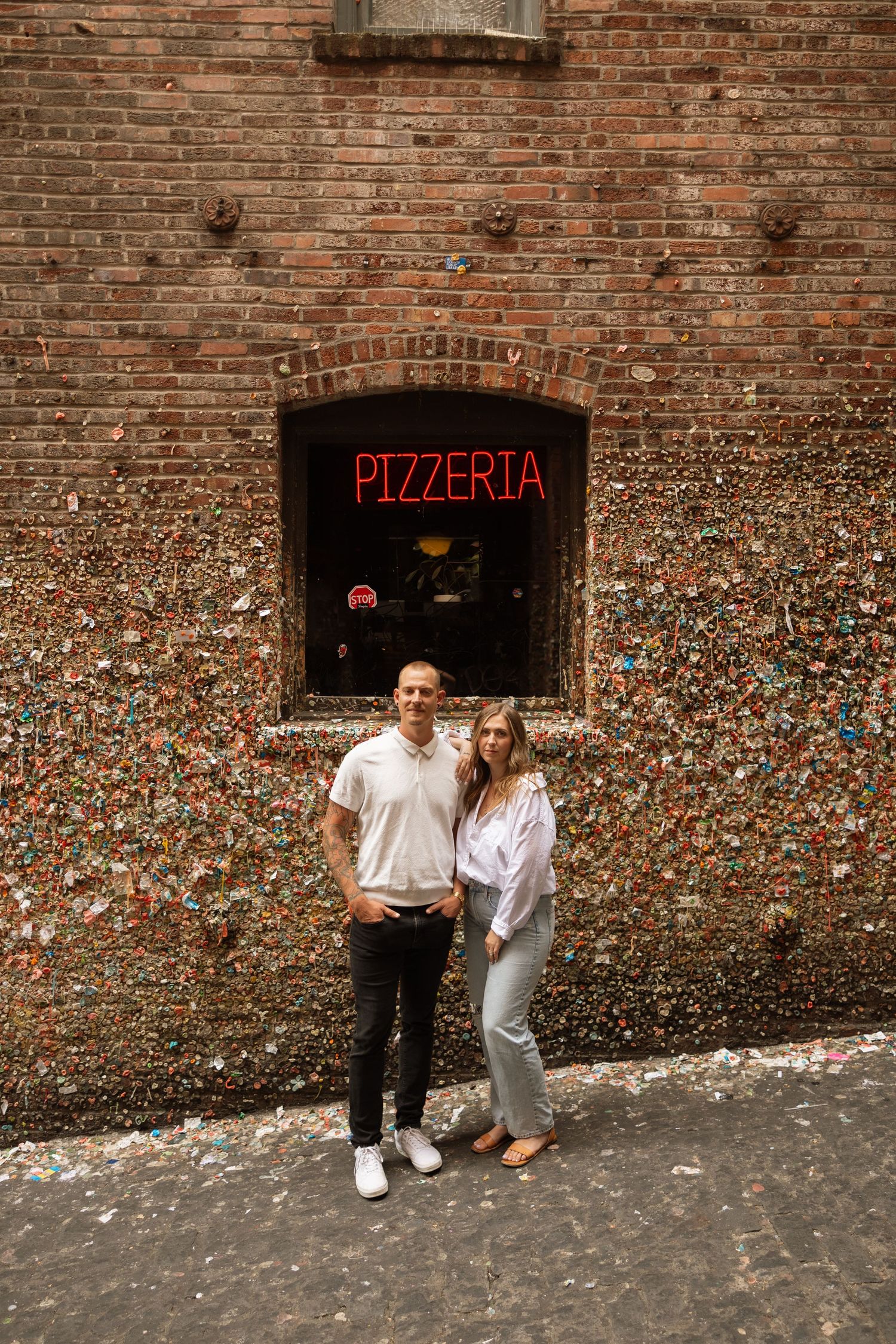 A couple poses in front of a brick wall with a neon pizzeria sign covered in colorful gum.