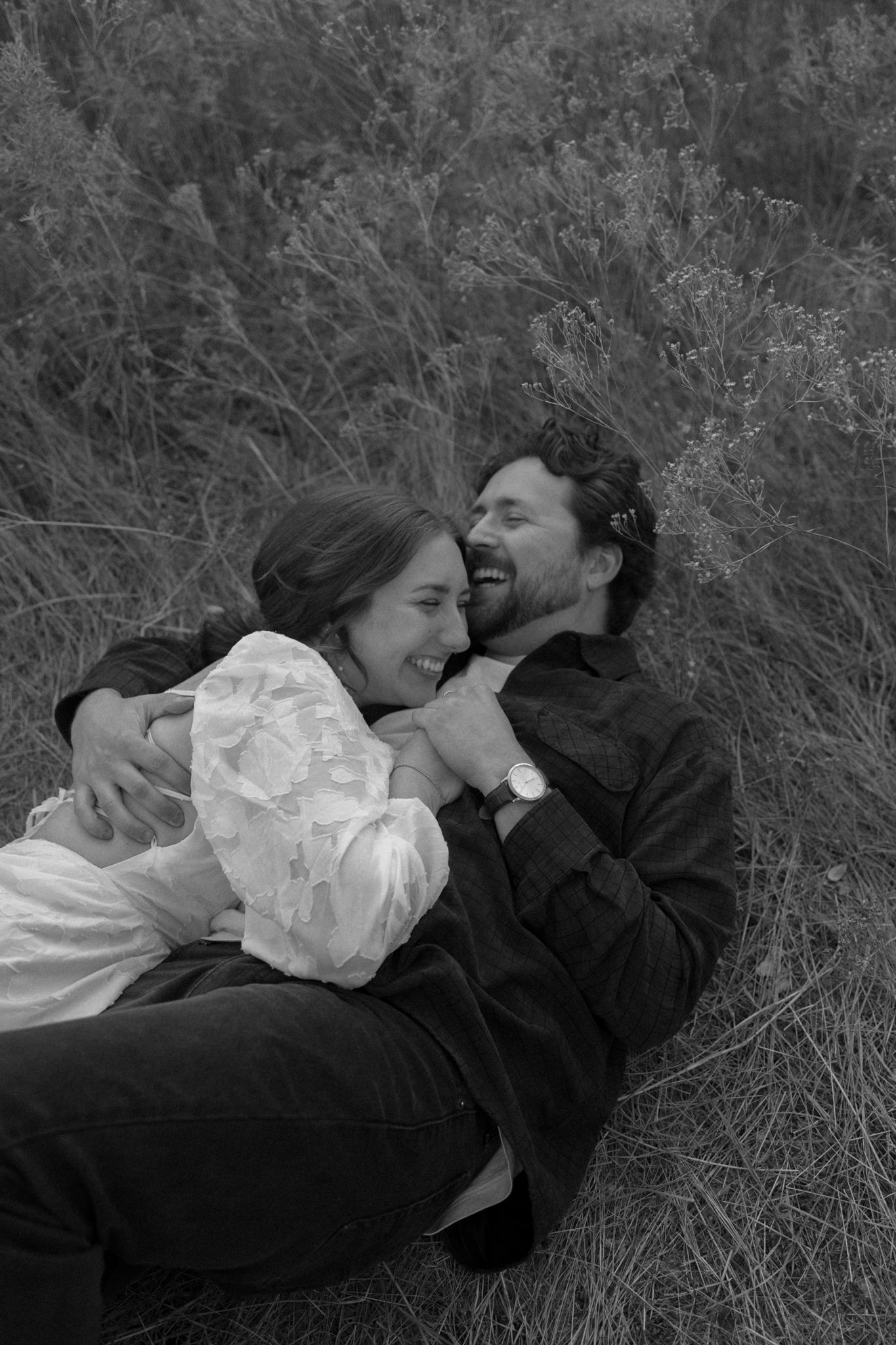 A black and white photo captures a tender moment between a couple lying in tall grass.