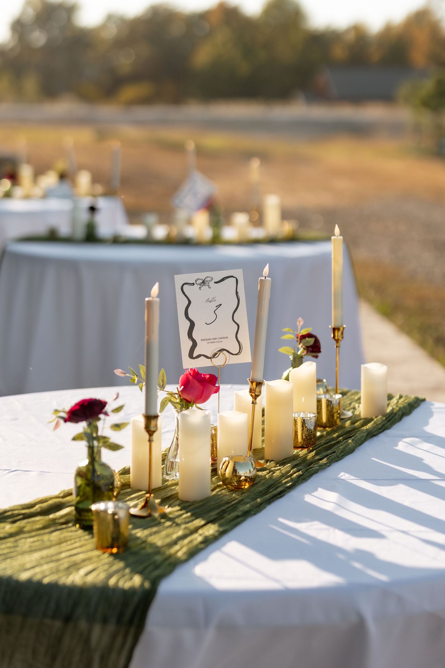An elegant wedding table setting with white candles and roses on a green runner for outdoor dining.