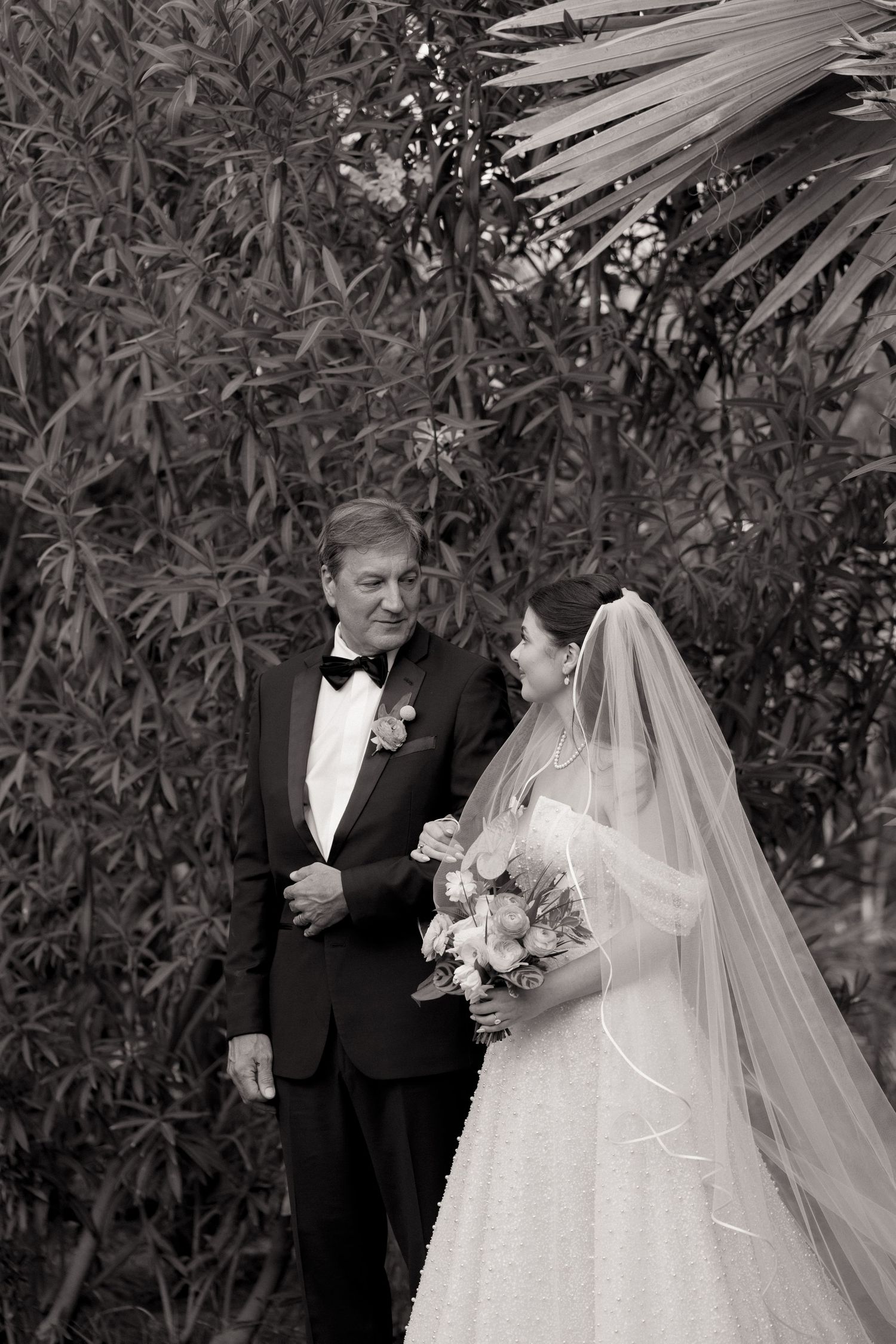 Black and white portrait of bride in veil holding bouquet standing beside older gentleman in tuxedo outdoors.