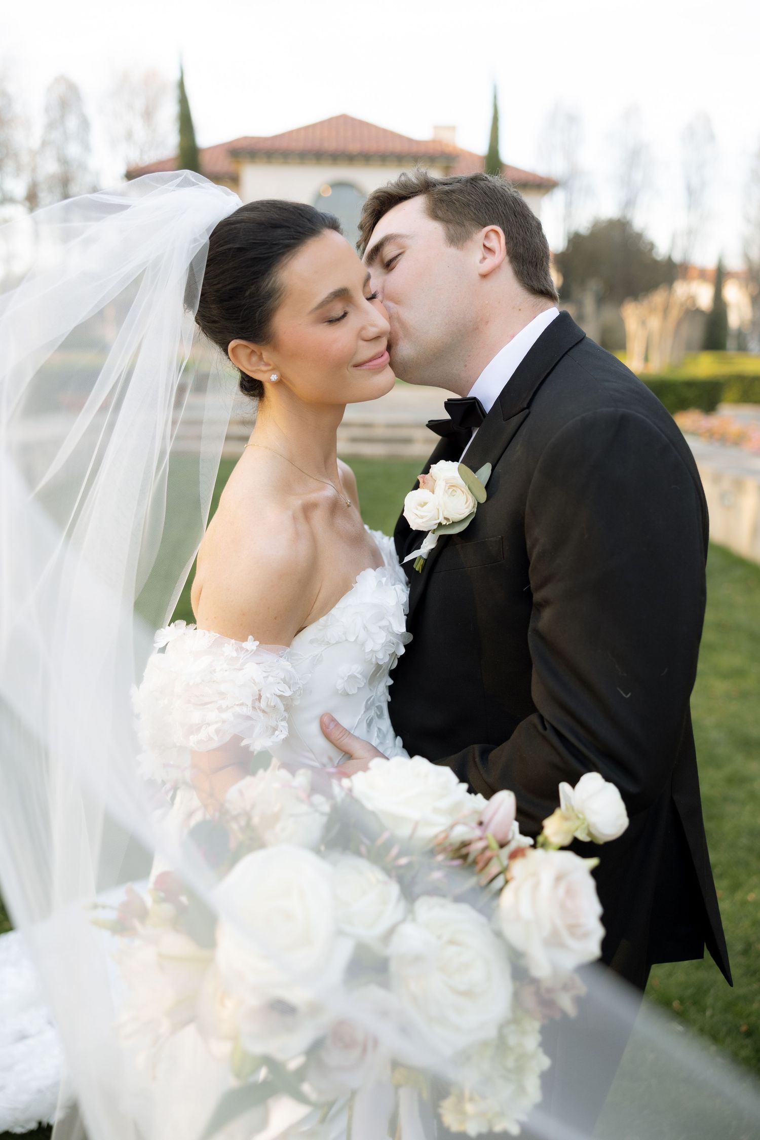 Groom kisses bride on cheek as her veil flows with white floral bouquet in foreground.