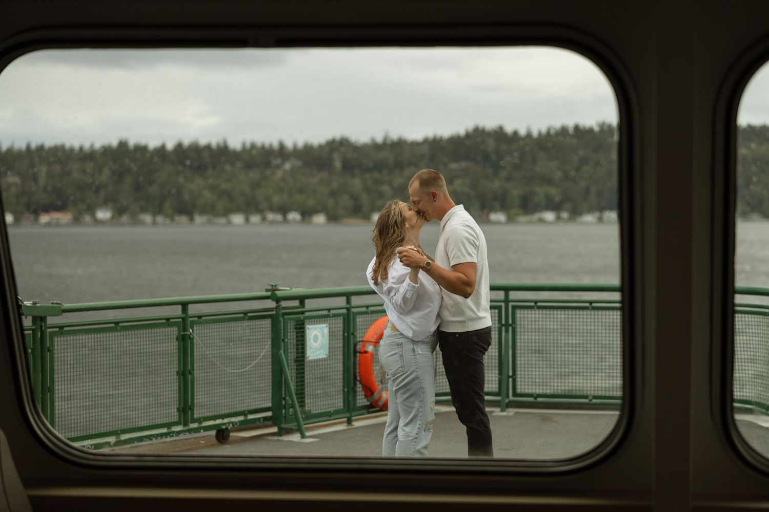 Couple sharing moment on ferry deck with water and forested shore in background.
