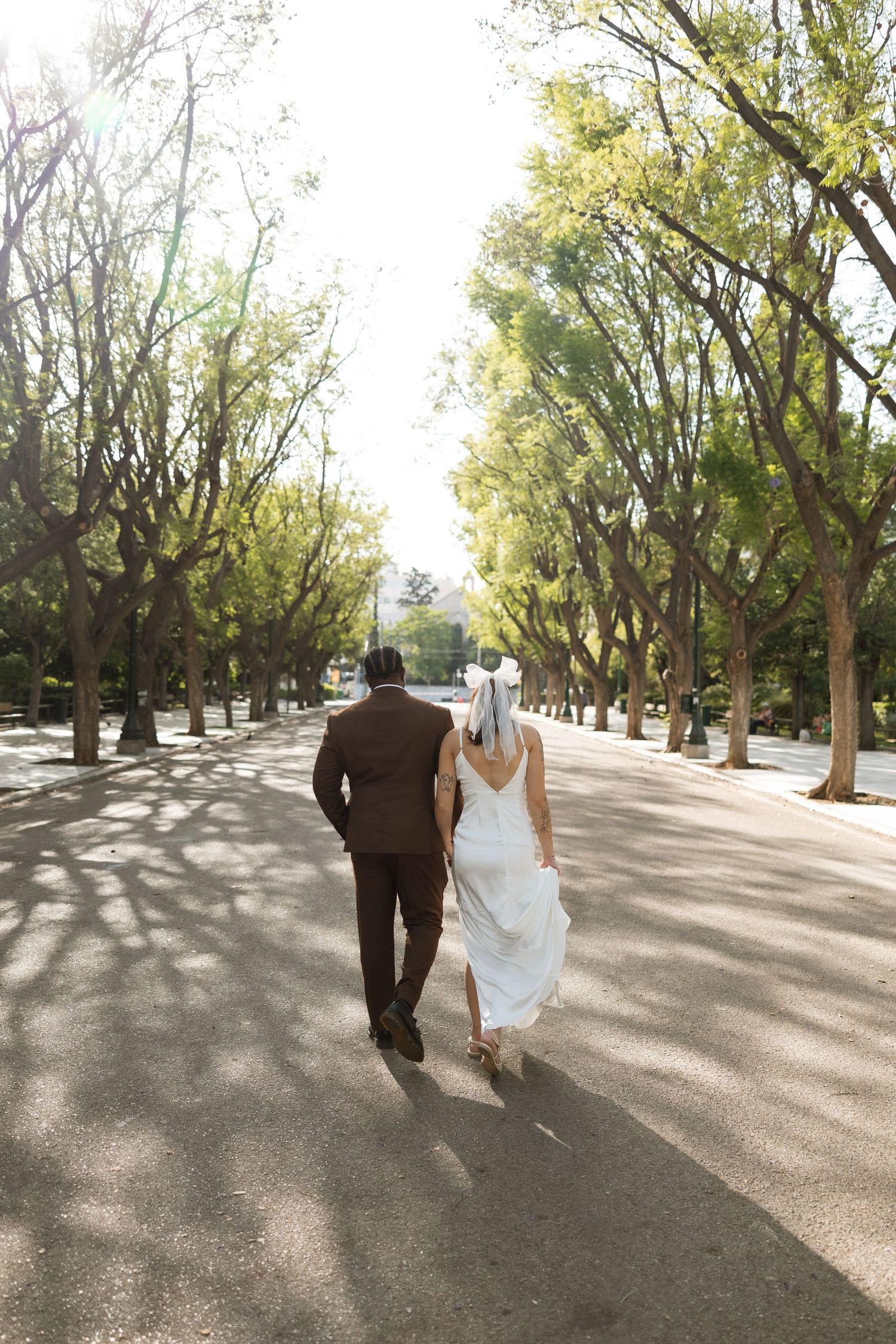 Wedding couple walks down tree-lined path with sunlight filtering through branches.