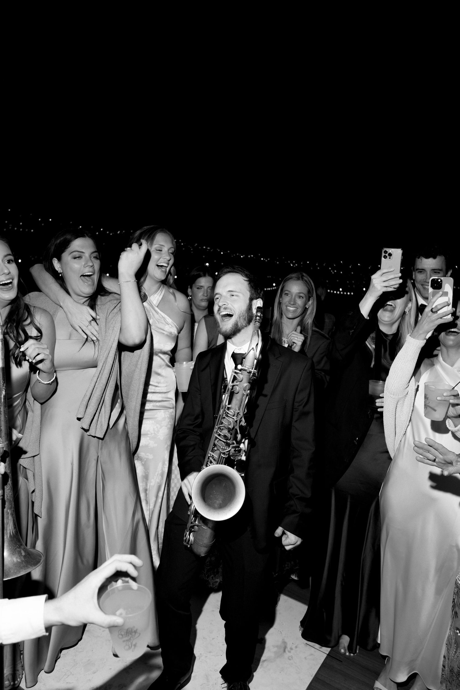 A saxophonist performs energetically among celebrating wedding guests at a nighttime outdoor reception in black and white.