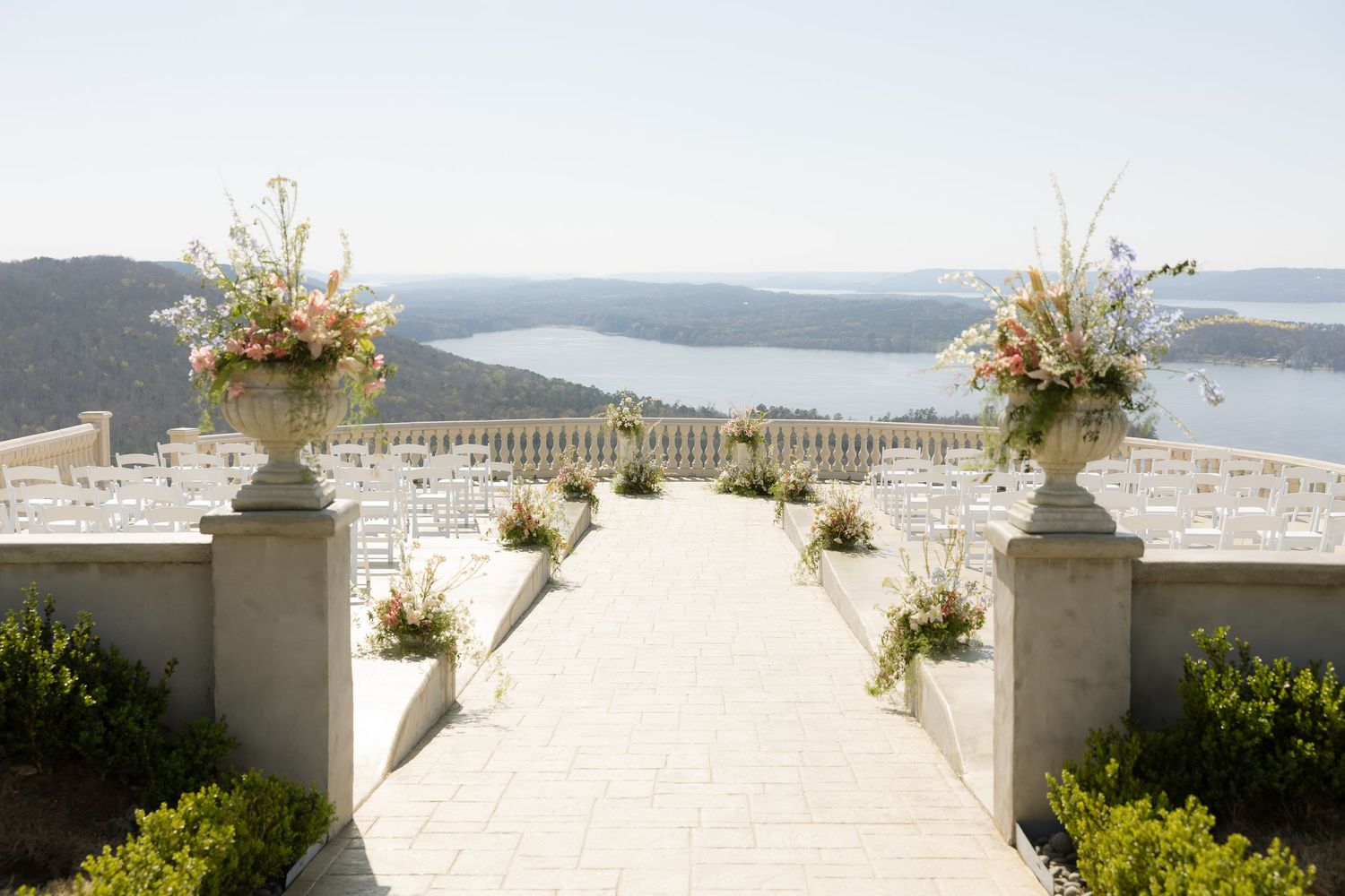 Elegant outdoor wedding ceremony aisle with floral urns overlooking a scenic lake and rolling hills.