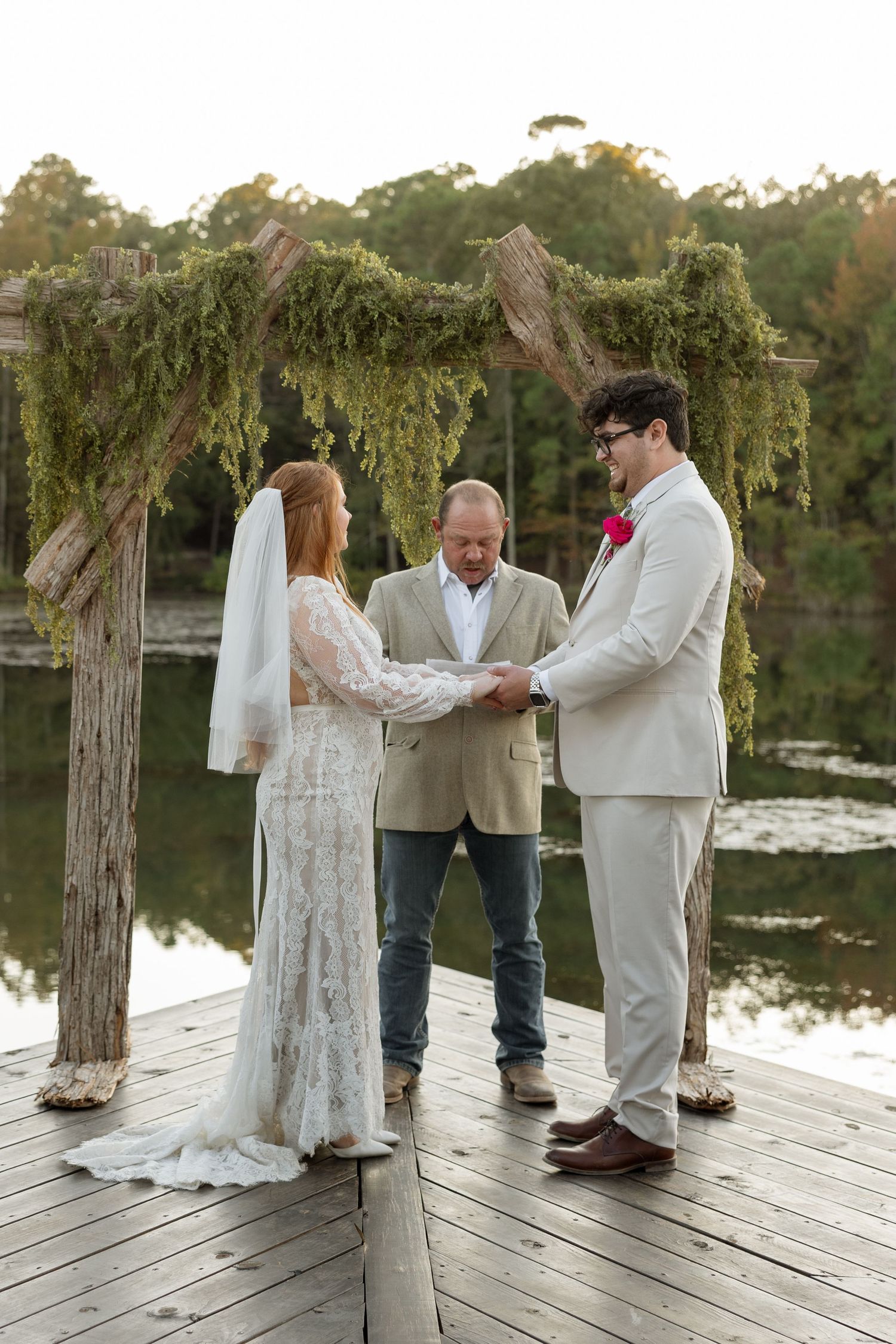 A wedding ceremony under a moss-covered arch on a wooden dock by the lake.