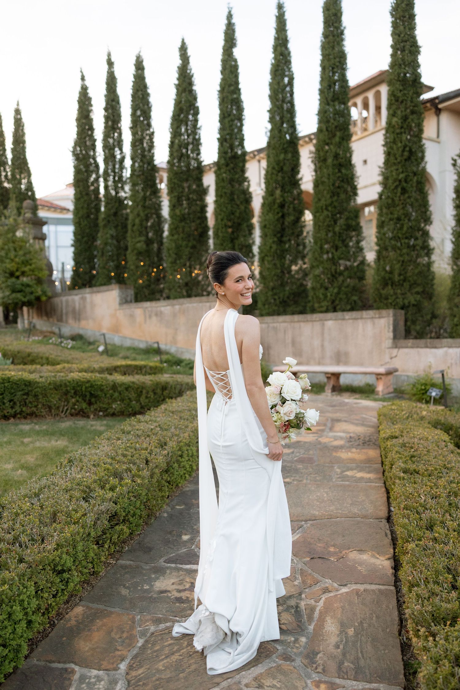 Bride in backless white gown smiling over shoulder along garden pathway lined with cypress trees.
