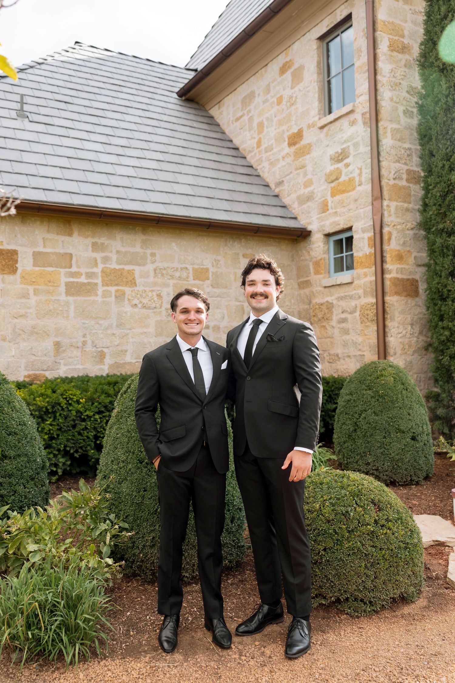 Two groomsmen in black suits pose together on a stone path outside an elegant stone building.