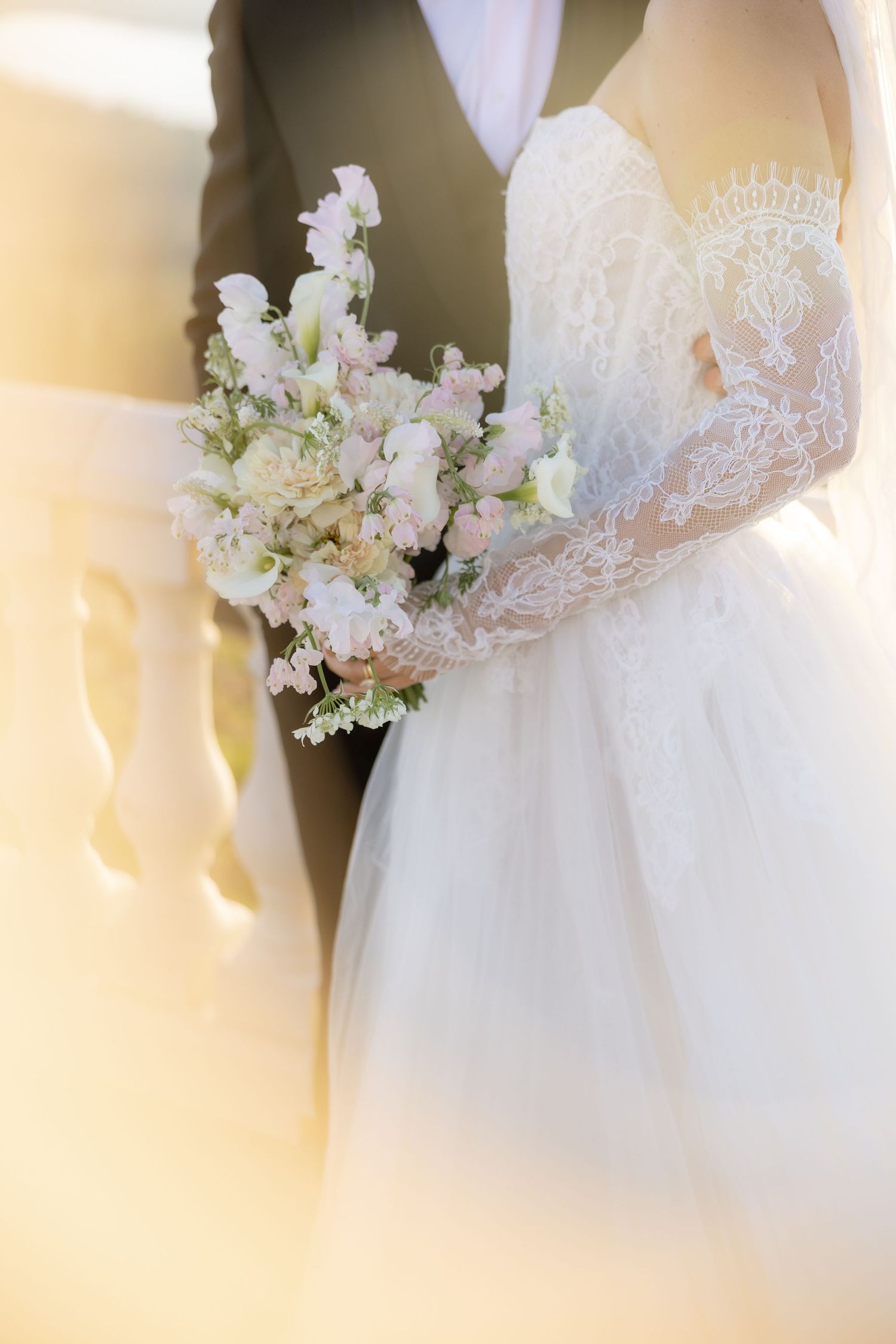 Bride in lace wedding gown holding a delicate bouquet of white and pink flowers beside groom.