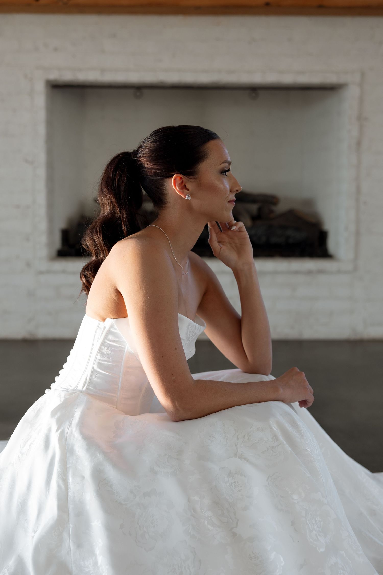 Elegant bride in white strapless gown sits pensively in front of white brick fireplace in romantic portrait.