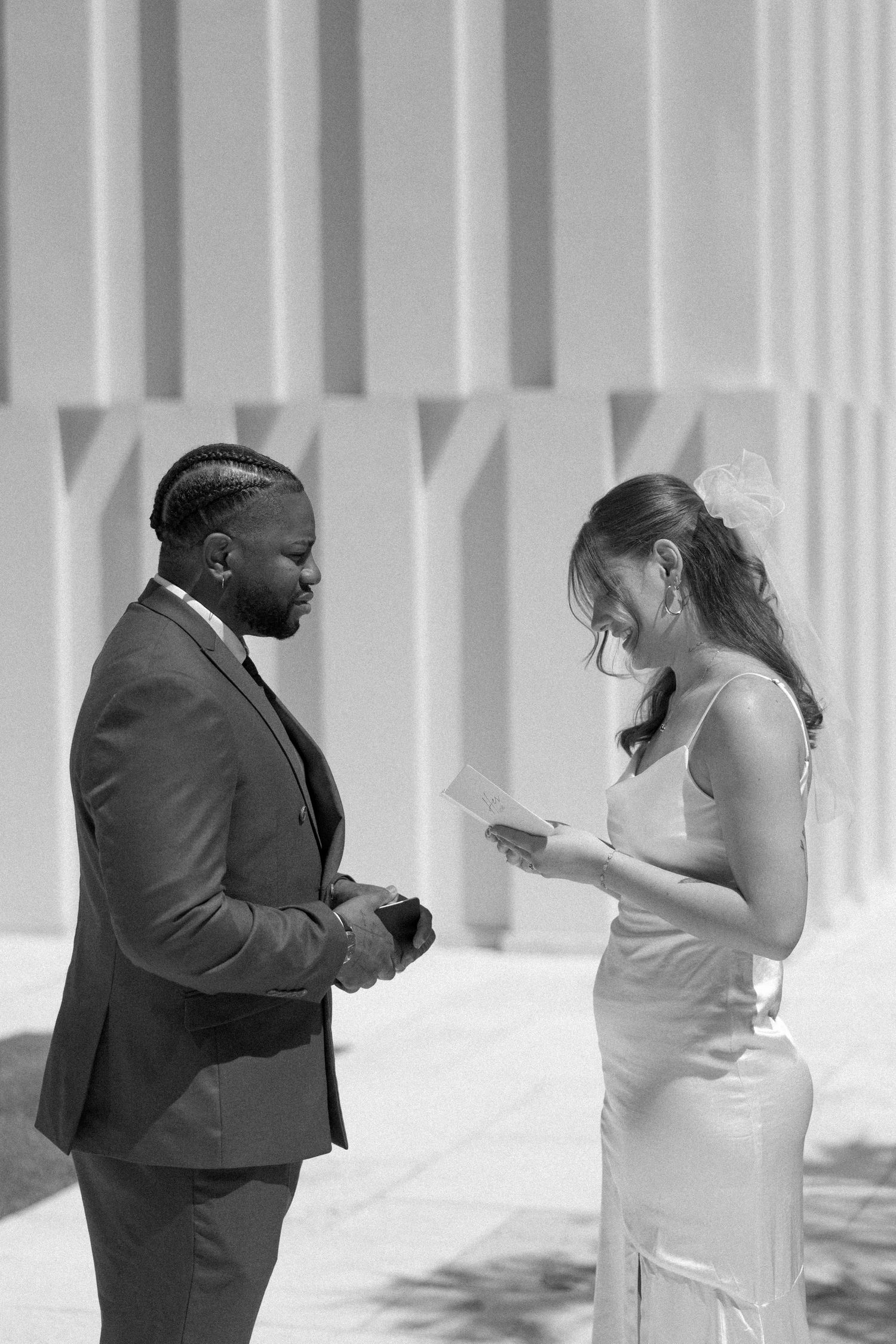 A black and white photo captures a couple exchanging vows outdoors by a modern building.