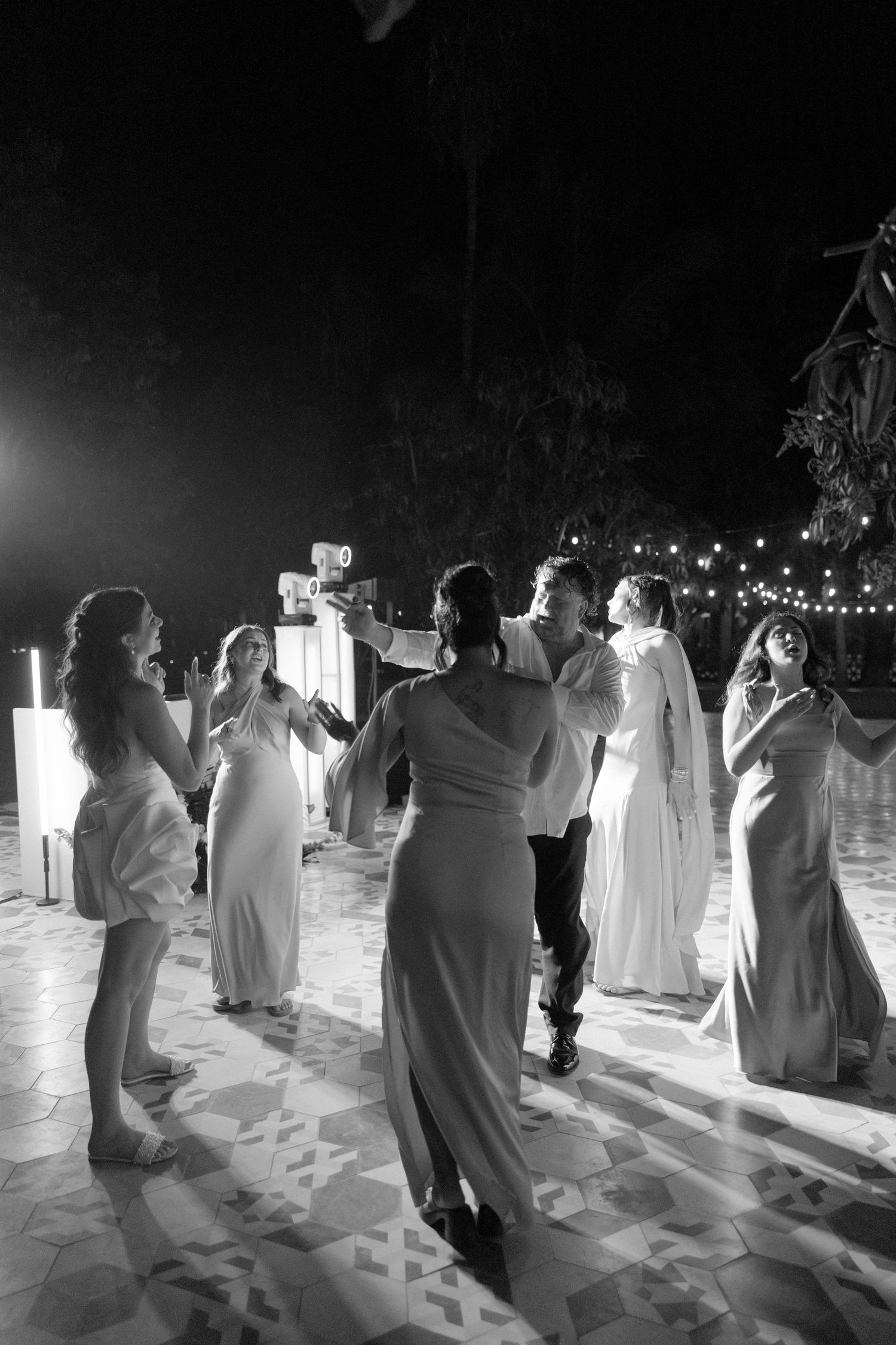 Black and white photo of wedding guests dancing together on a patterned outdoor dance floor at night.