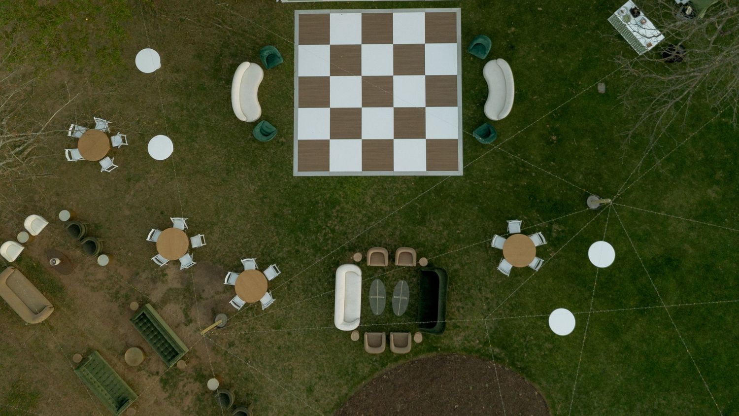 Aerial view of a giant outdoor chessboard surrounded by garden furniture on a green lawn.