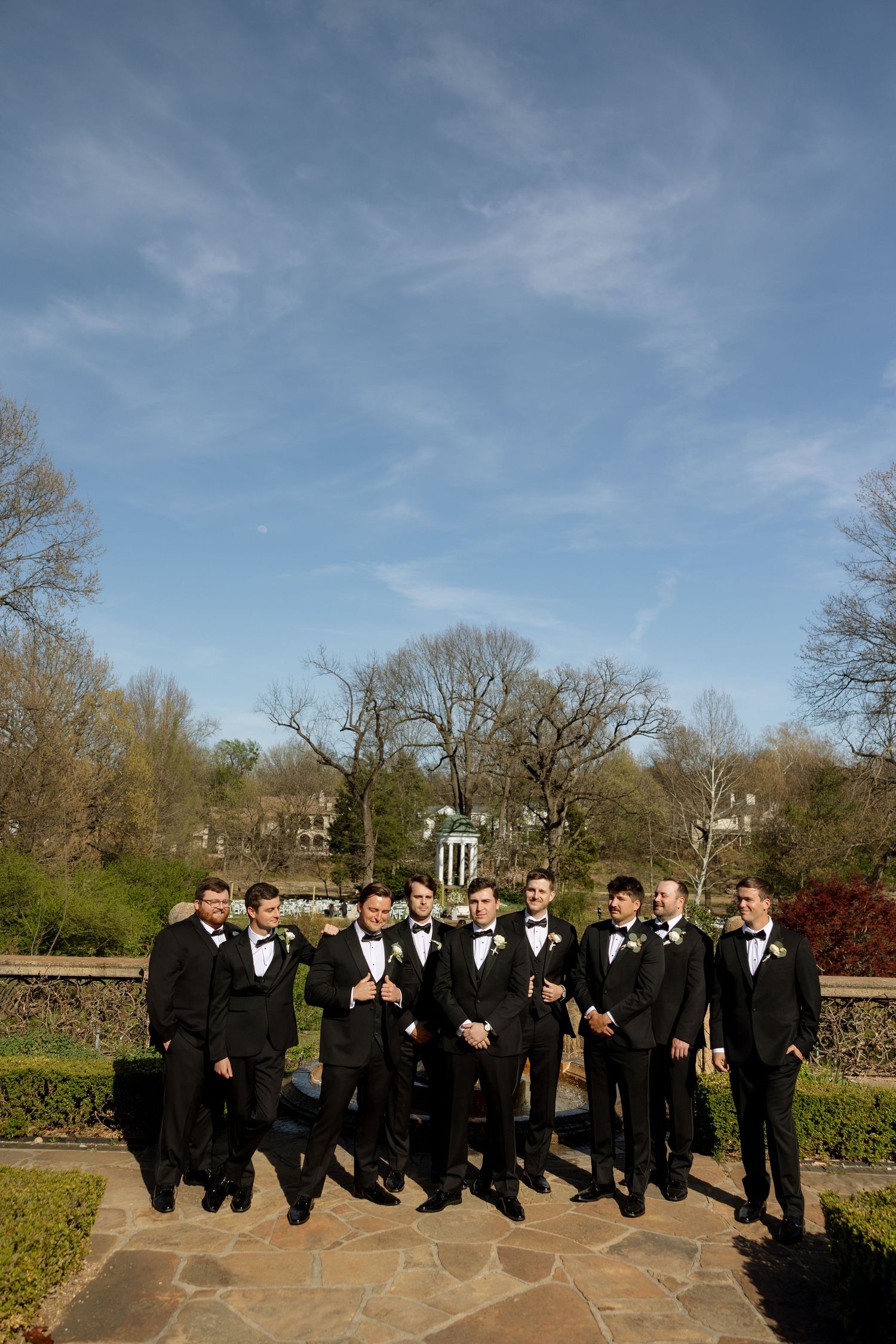 Groomsmen in black tuxedos posing on stone patio with blue sky and garden backdrop.