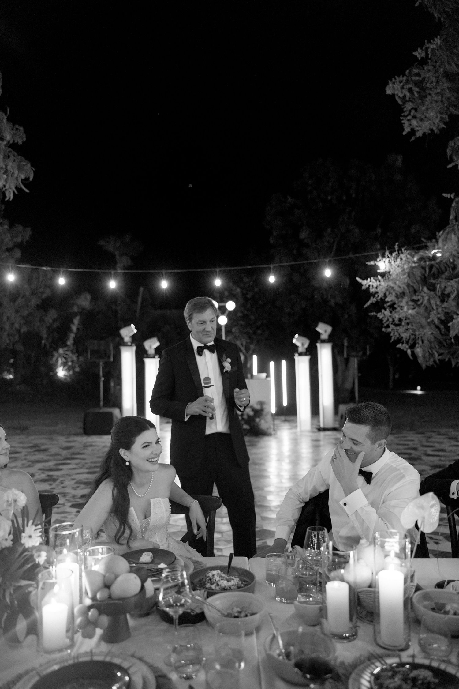 Bride and groom laugh during a wedding reception speech under string lights at a candlelit outdoor dinner table.