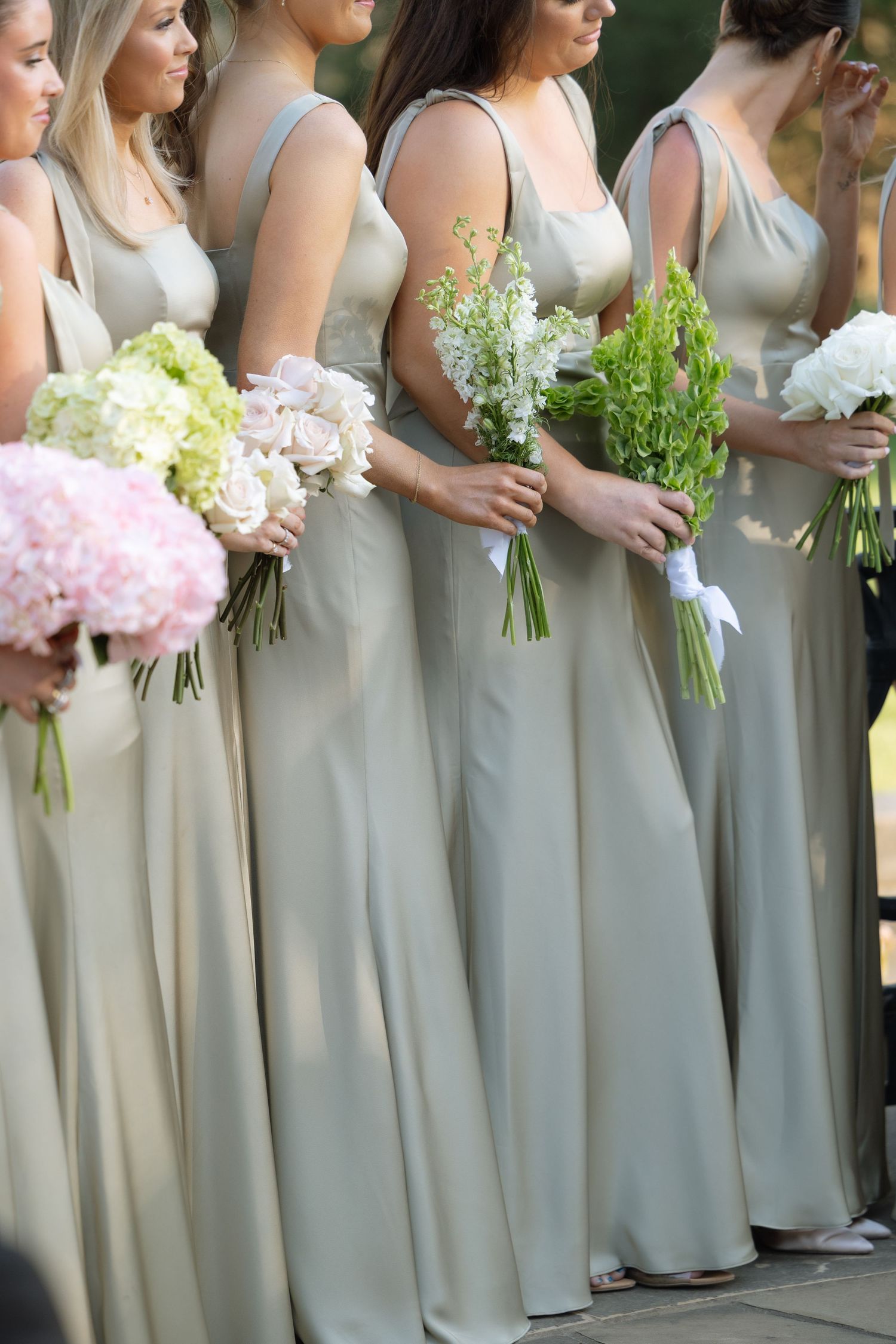 Row of bridesmaids in sage green gowns holding varied floral bouquets of pink, white, and green blooms.