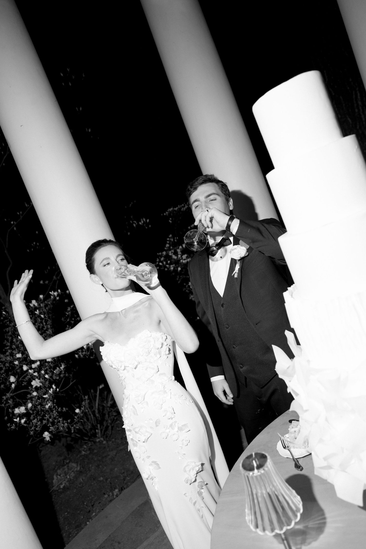 Bride and groom toasting with champagne beside their wedding cake in black and white.