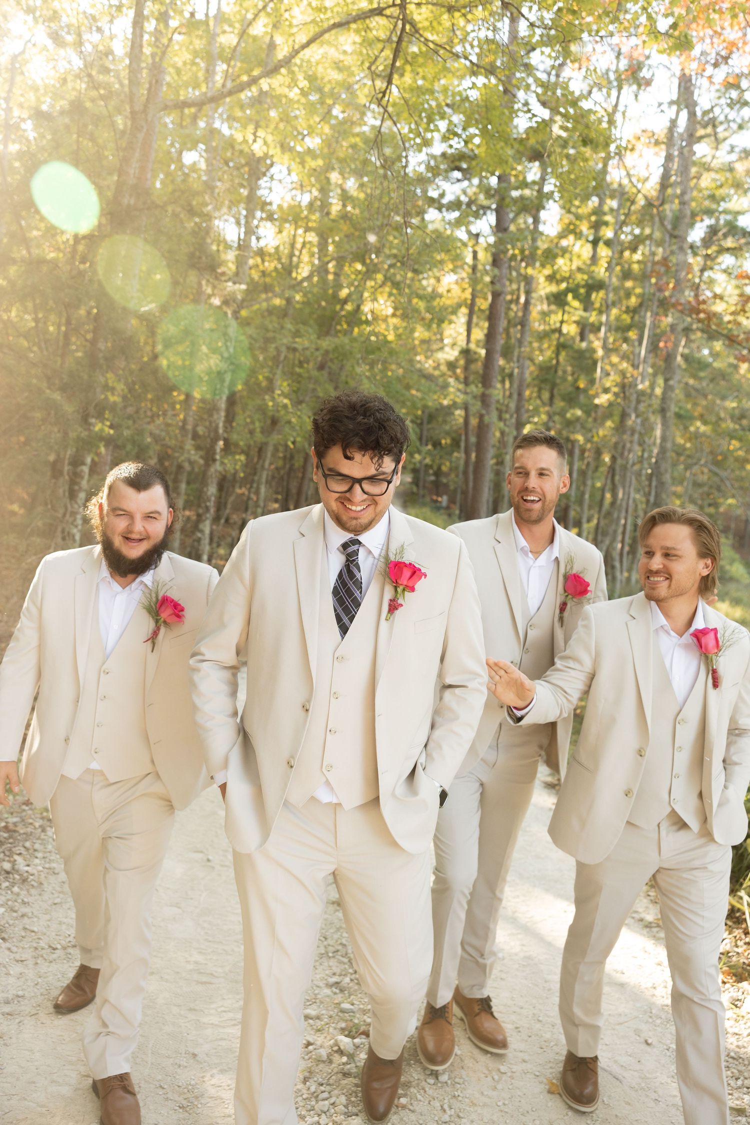 Sunlit portrait of groomsmen in cream suits laughing together on a wooded path.