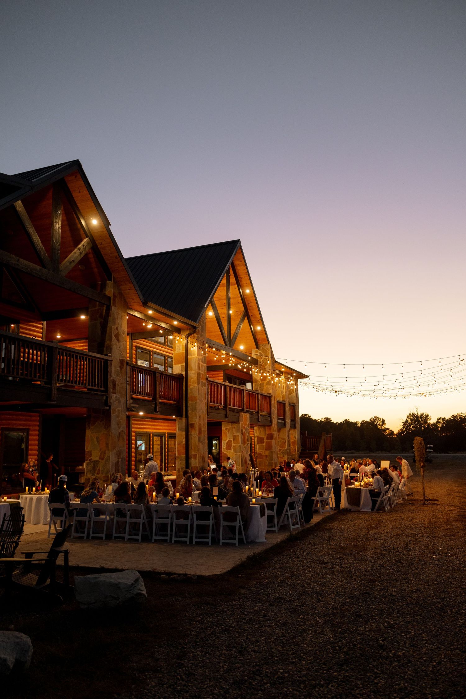 Wedding reception guests dine outside a rustic lodge at twilight under glowing string lights.