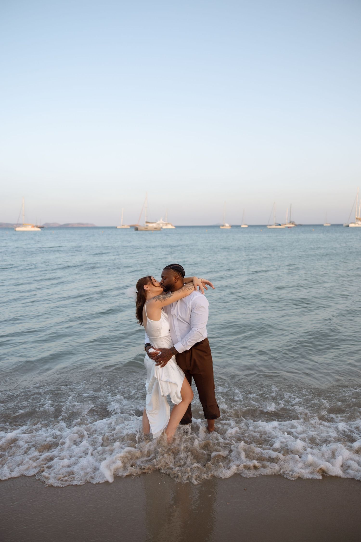 A romantic embrace in the ocean waves with sailboats dotting the horizon during golden hour.