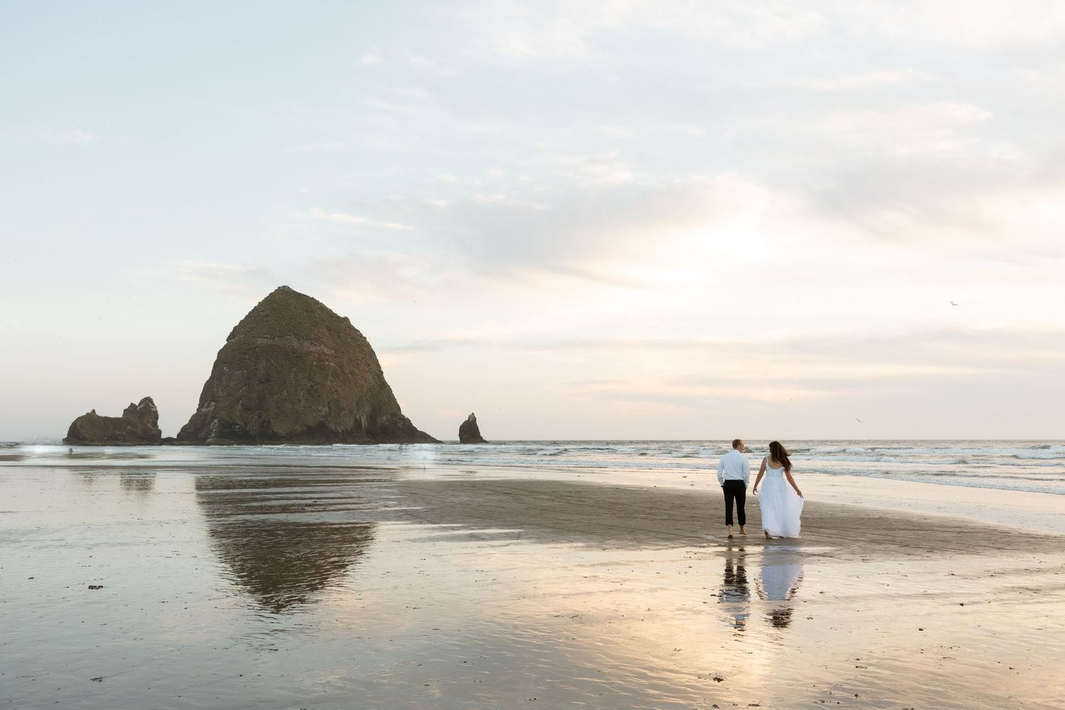 Couple in wedding attire stands on beach with iconic Haystack Rock at sunset.