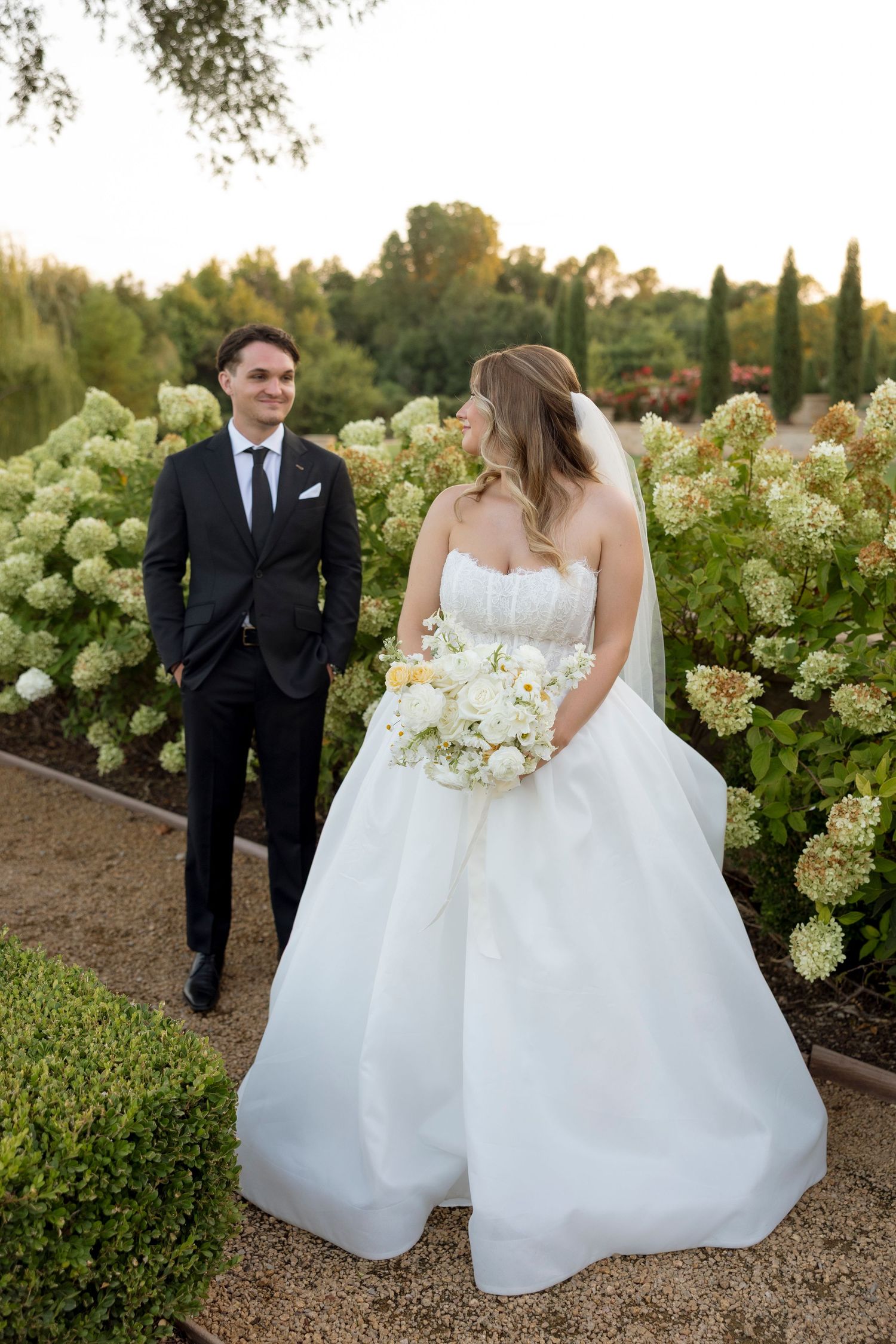 Wedding couple strolls through blooming hydrangea garden during sunset photoshoot.