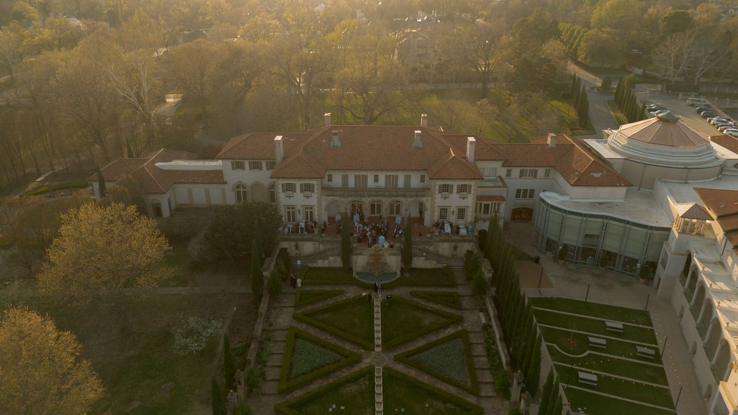 Aerial drone view of grand estate wedding venue surrounded by manicured formal gardens at sunset.