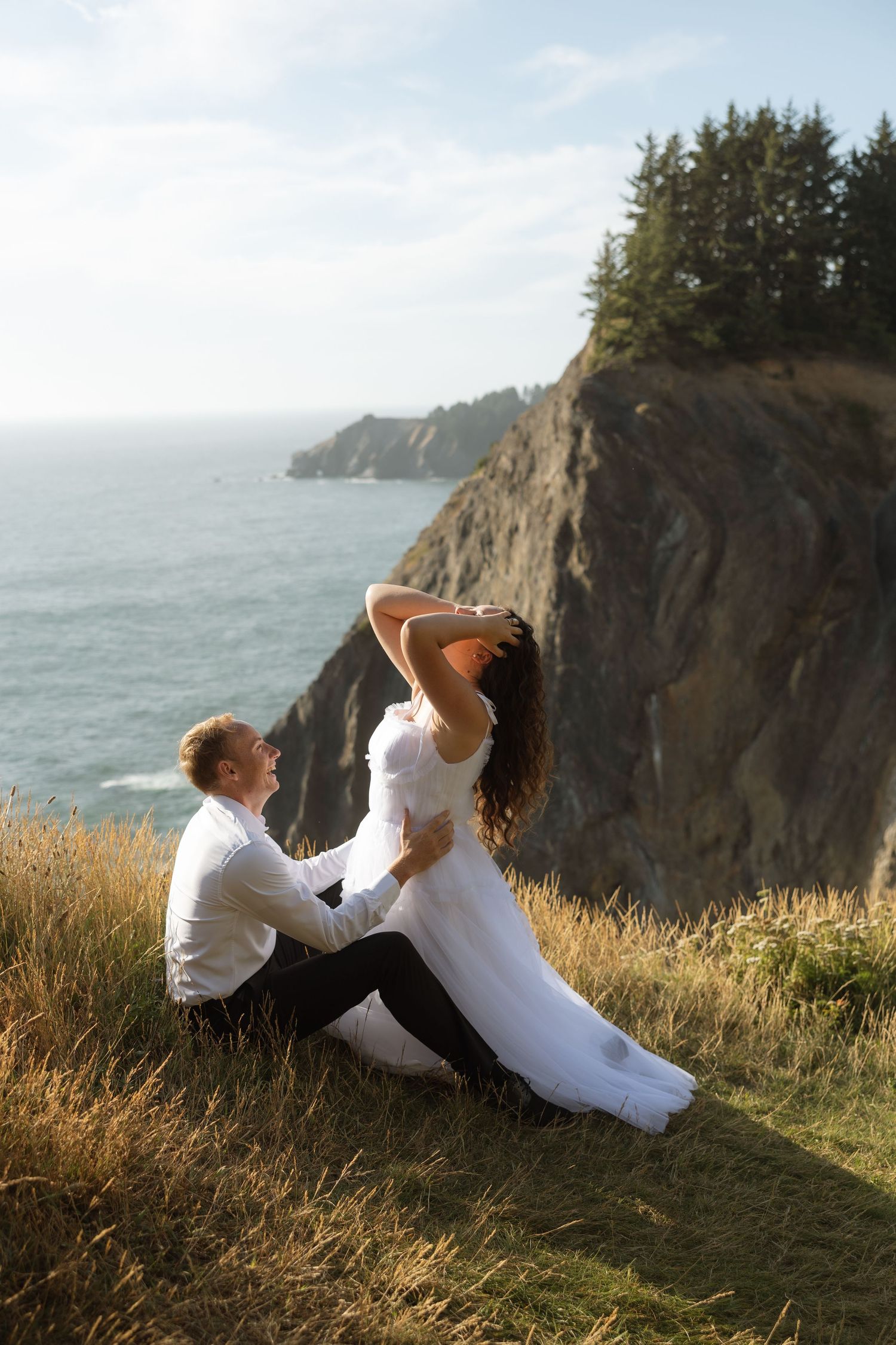 A couple shares a romantic moment on a grassy cliff overlooking the Pacific Ocean coastline.