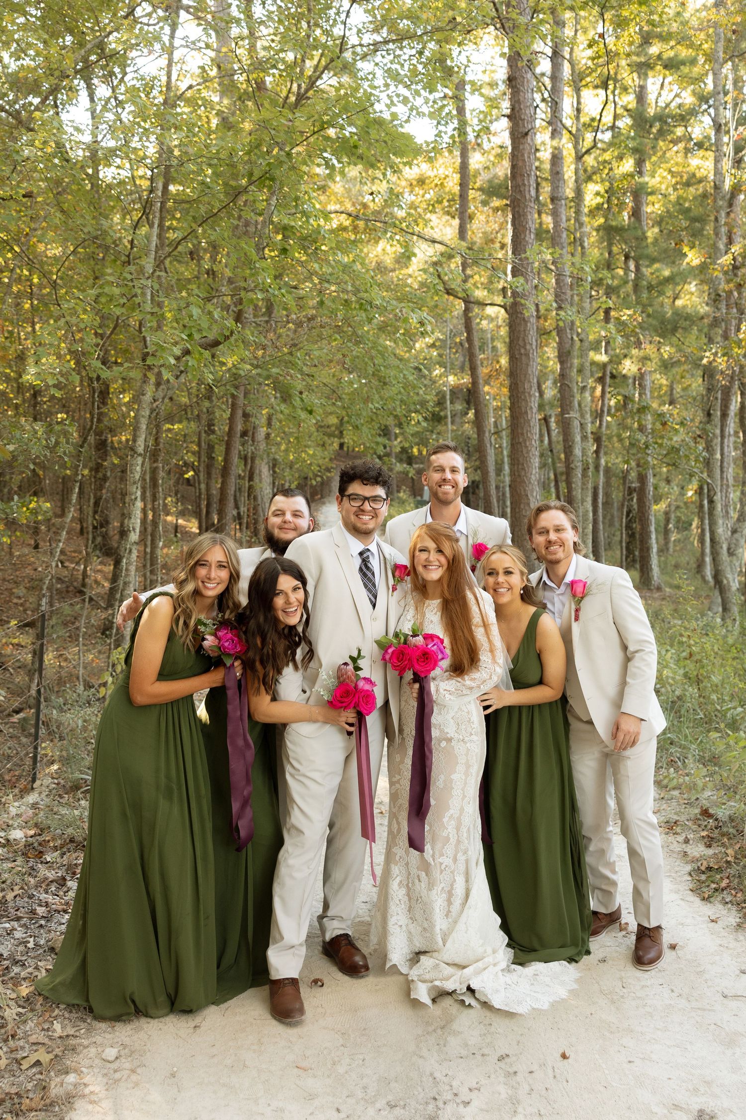 Wedding group portrait on forest path with bridesmaids in green dresses and groomsmen in light suits.