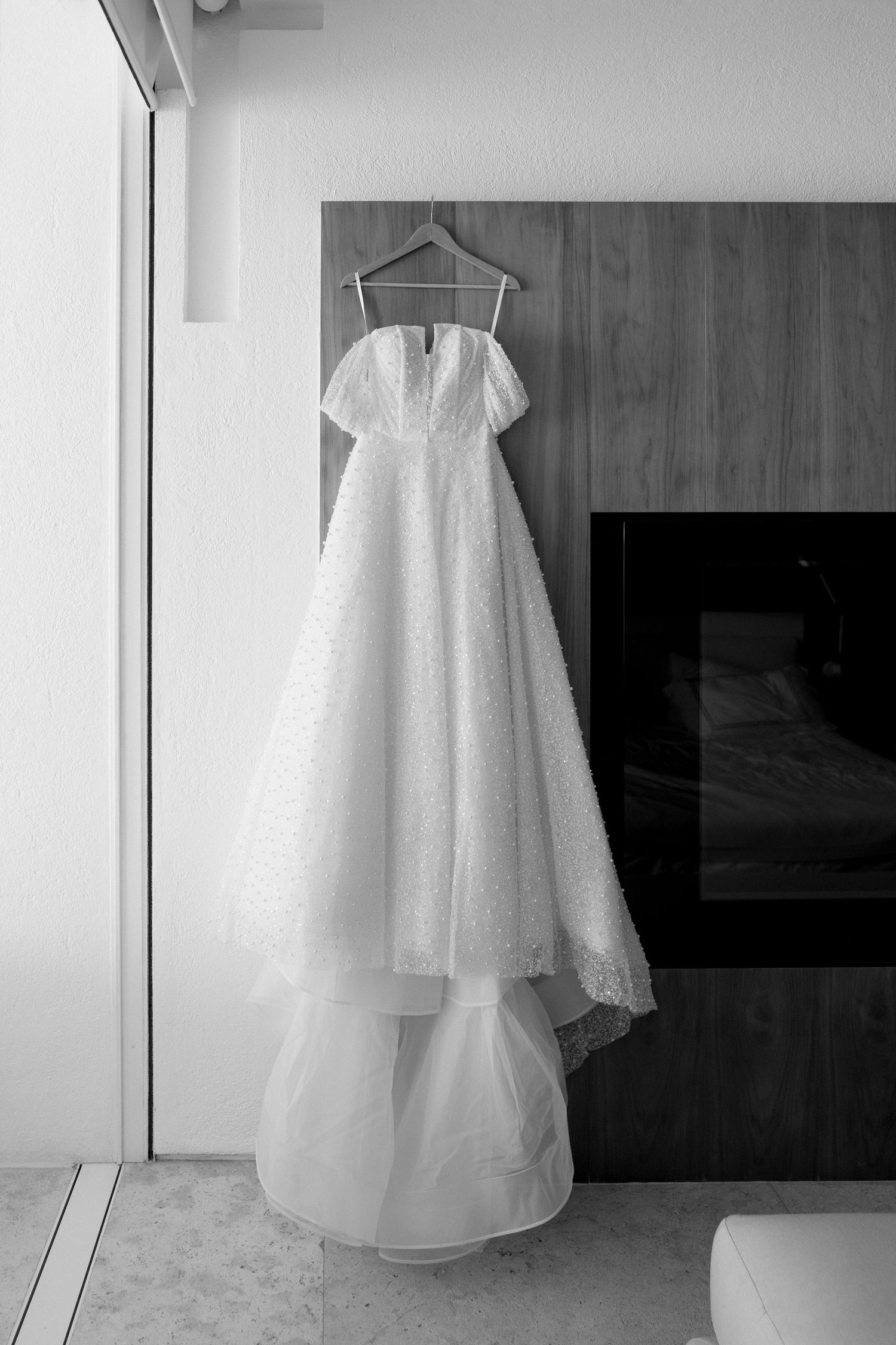 An elegant lace wedding dress hangs on a hanger against a modern interior backdrop in black and white.