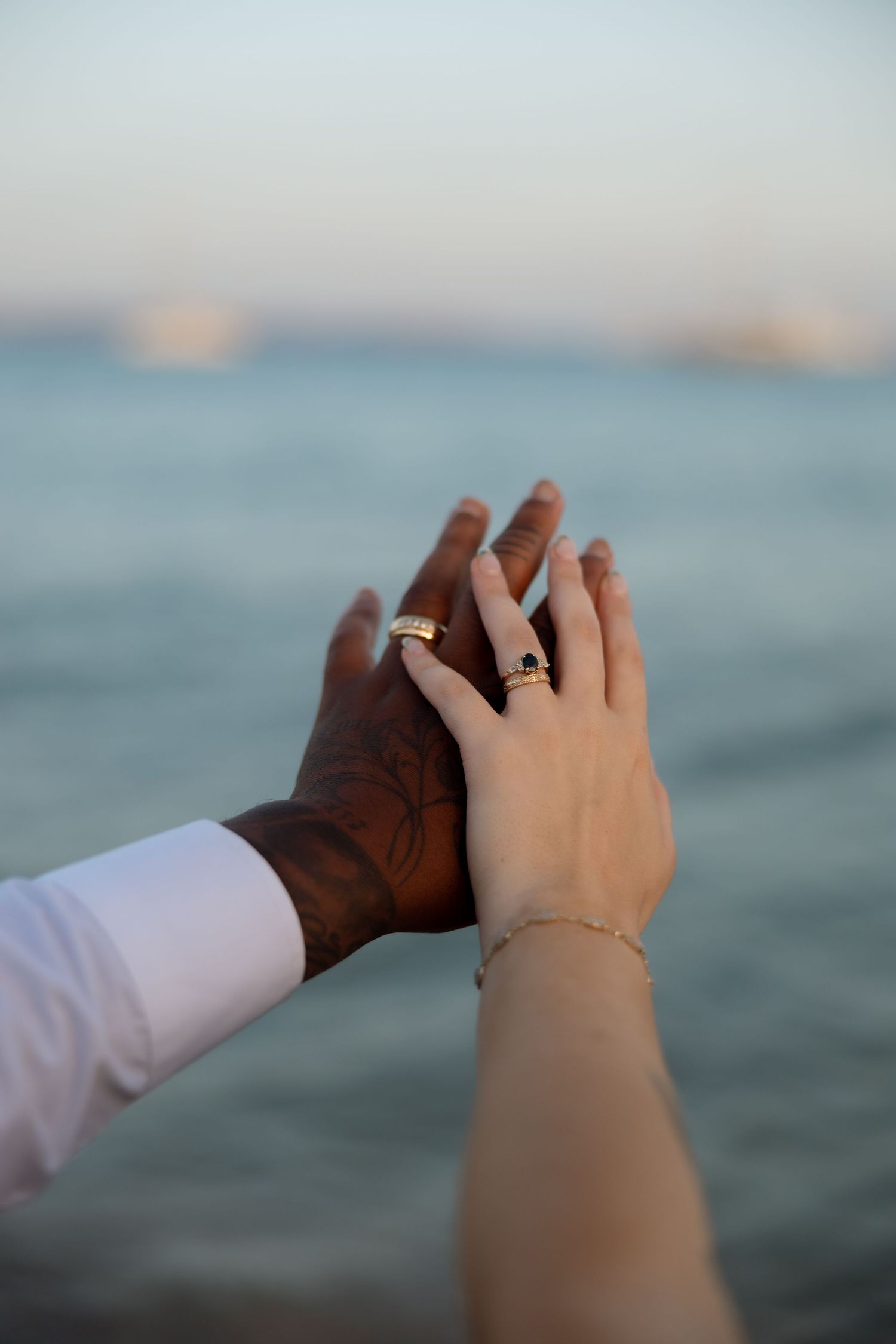Two hands wearing wedding rings intertwined against a blurred ocean backdrop.