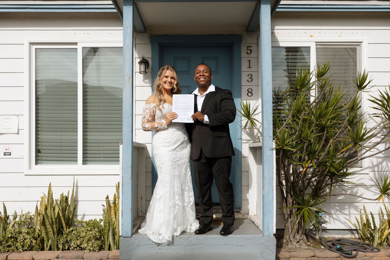 A wedding couple stands together in front of a white house entrance with palm plants visible.