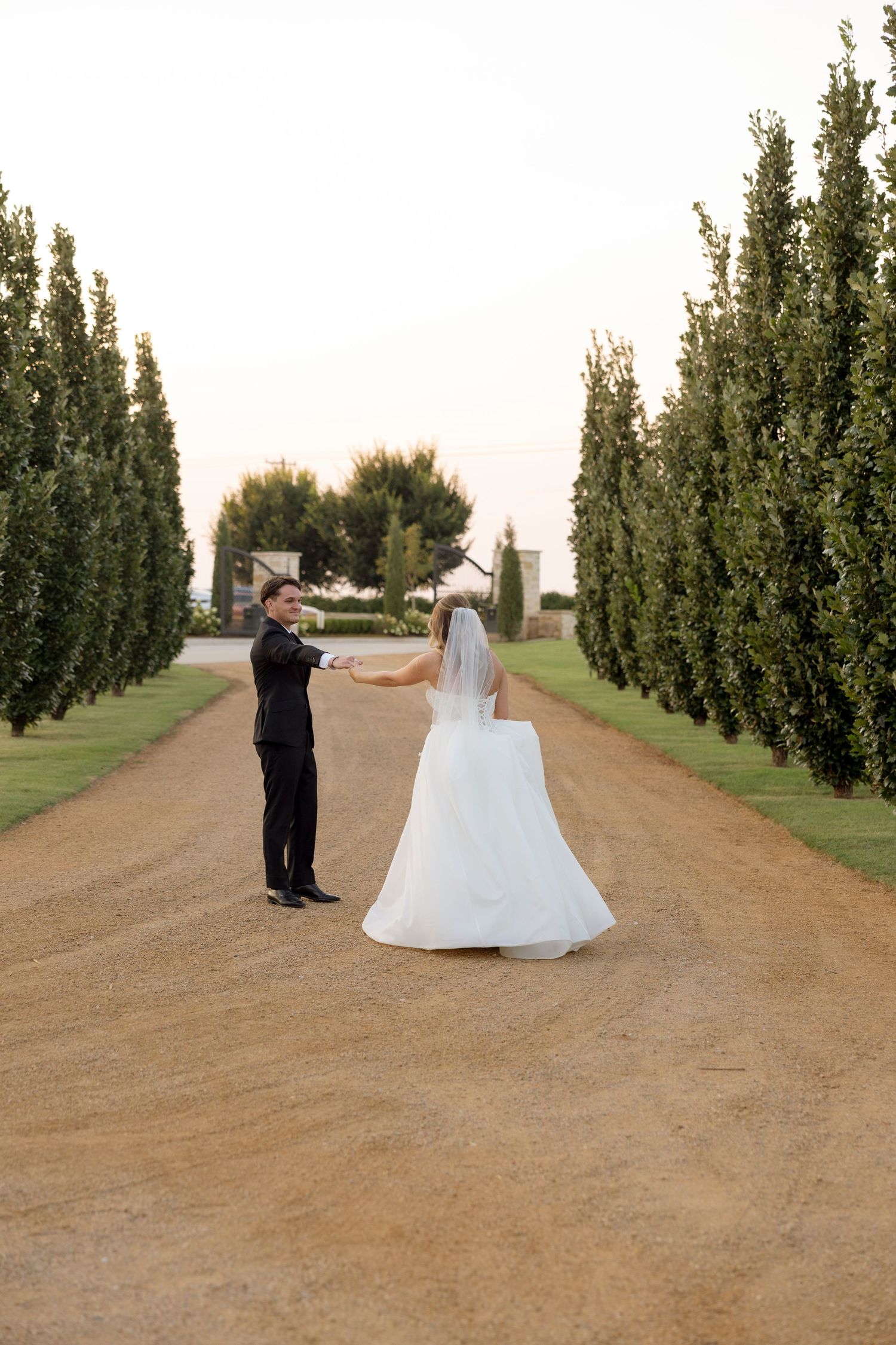 A newlywed couple dances together on a rustic dirt road lined with tall cypress trees.