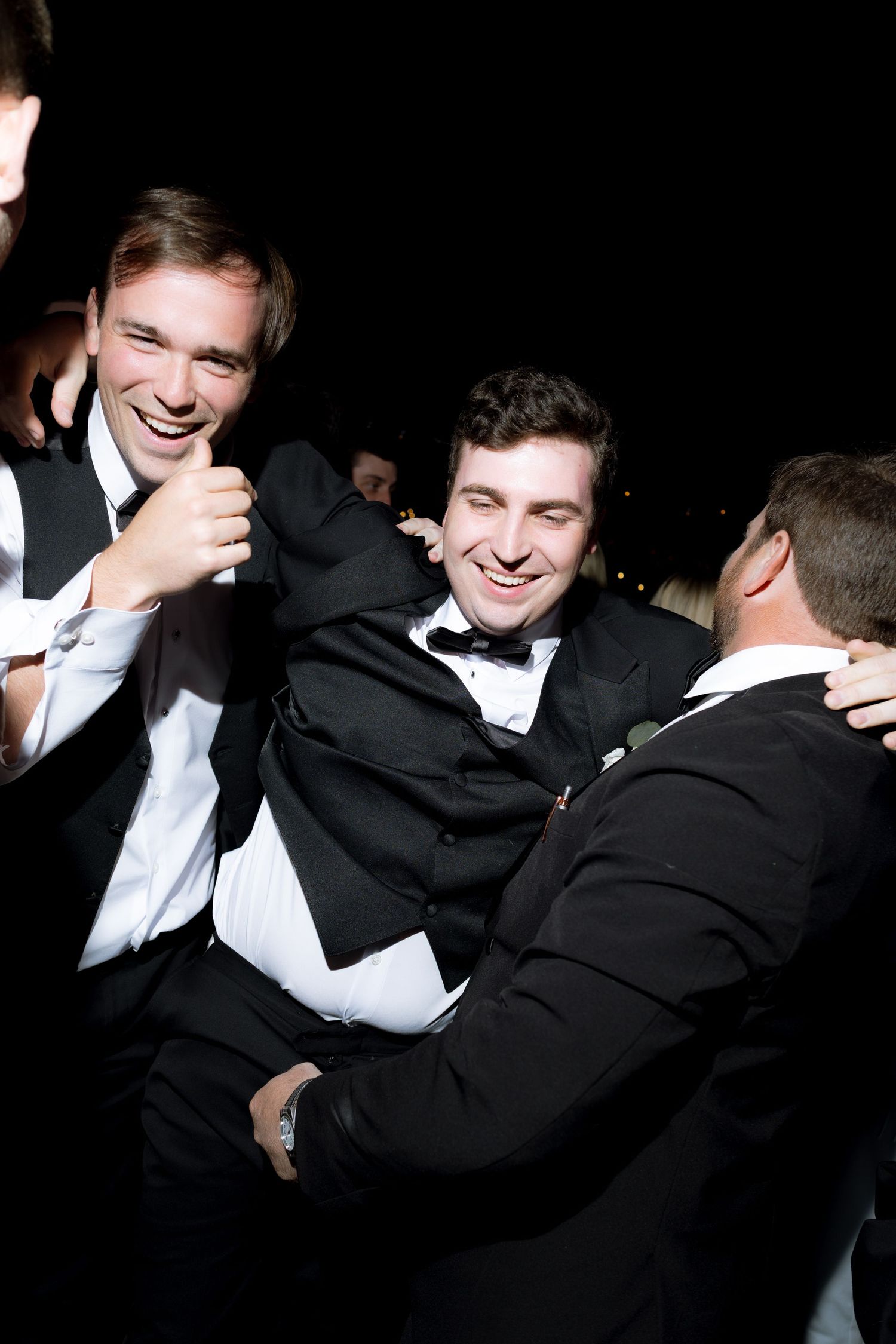 Groomsmen in black tuxedos laugh and embrace joyfully during a lively wedding reception celebration.