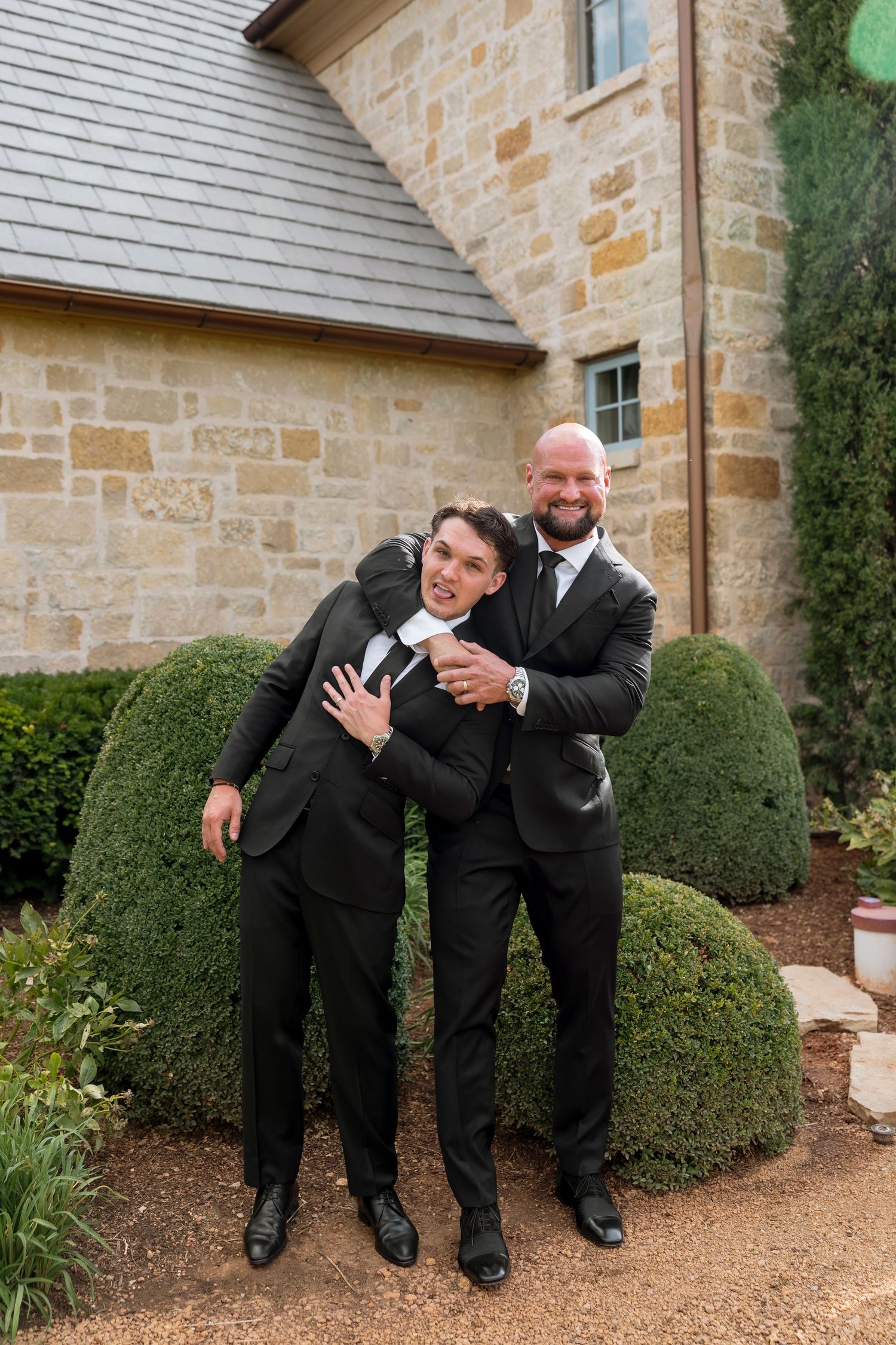 Two men in black tuxedos pose playfully in front of stone building with manicured shrubs.