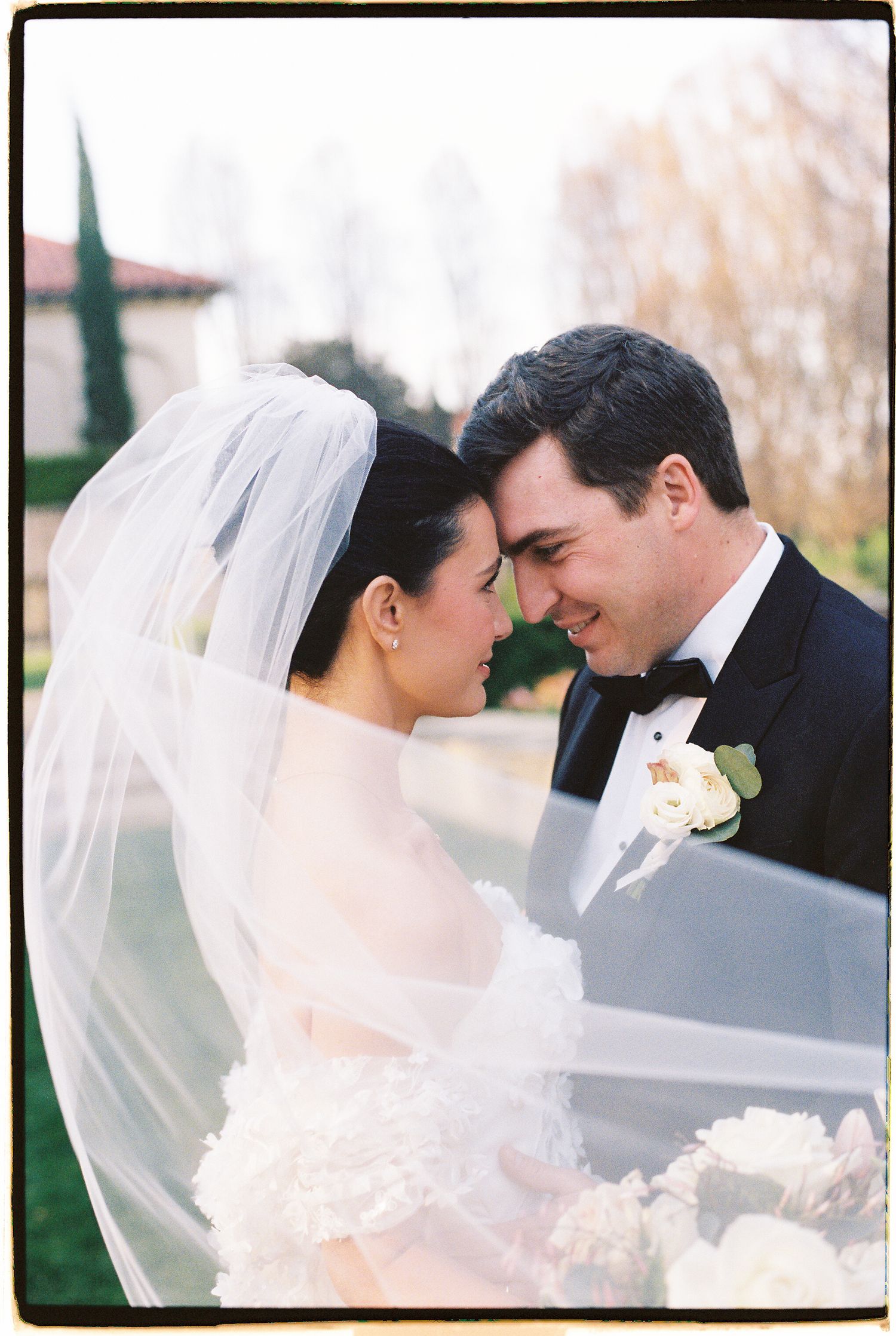Bride and groom sharing an intimate moment, bride wearing veil and groom in black tuxedo with white boutonniere.