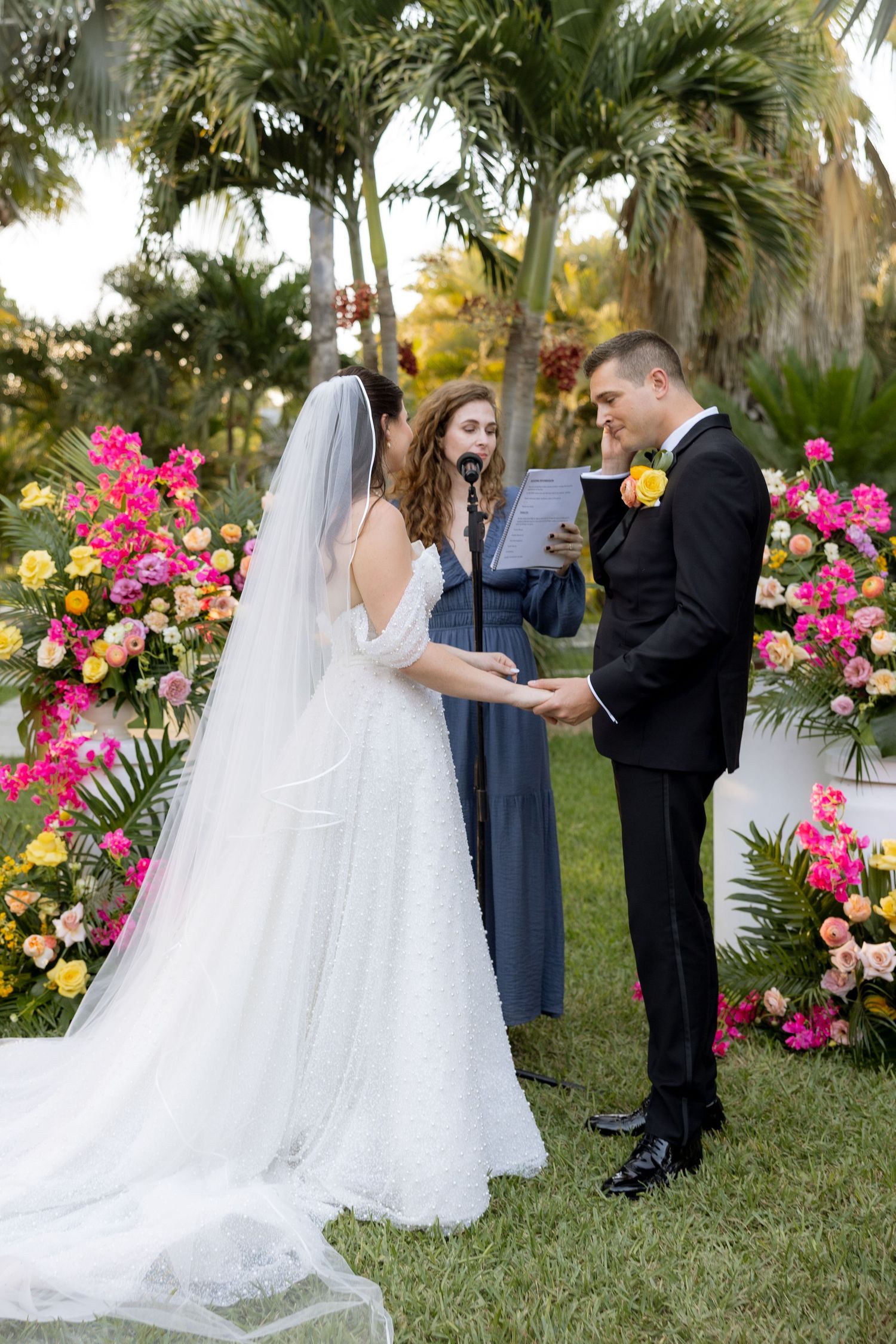 Bride and groom exchange vows outdoors surrounded by vibrant pink and yellow floral arrangements with an officiant.