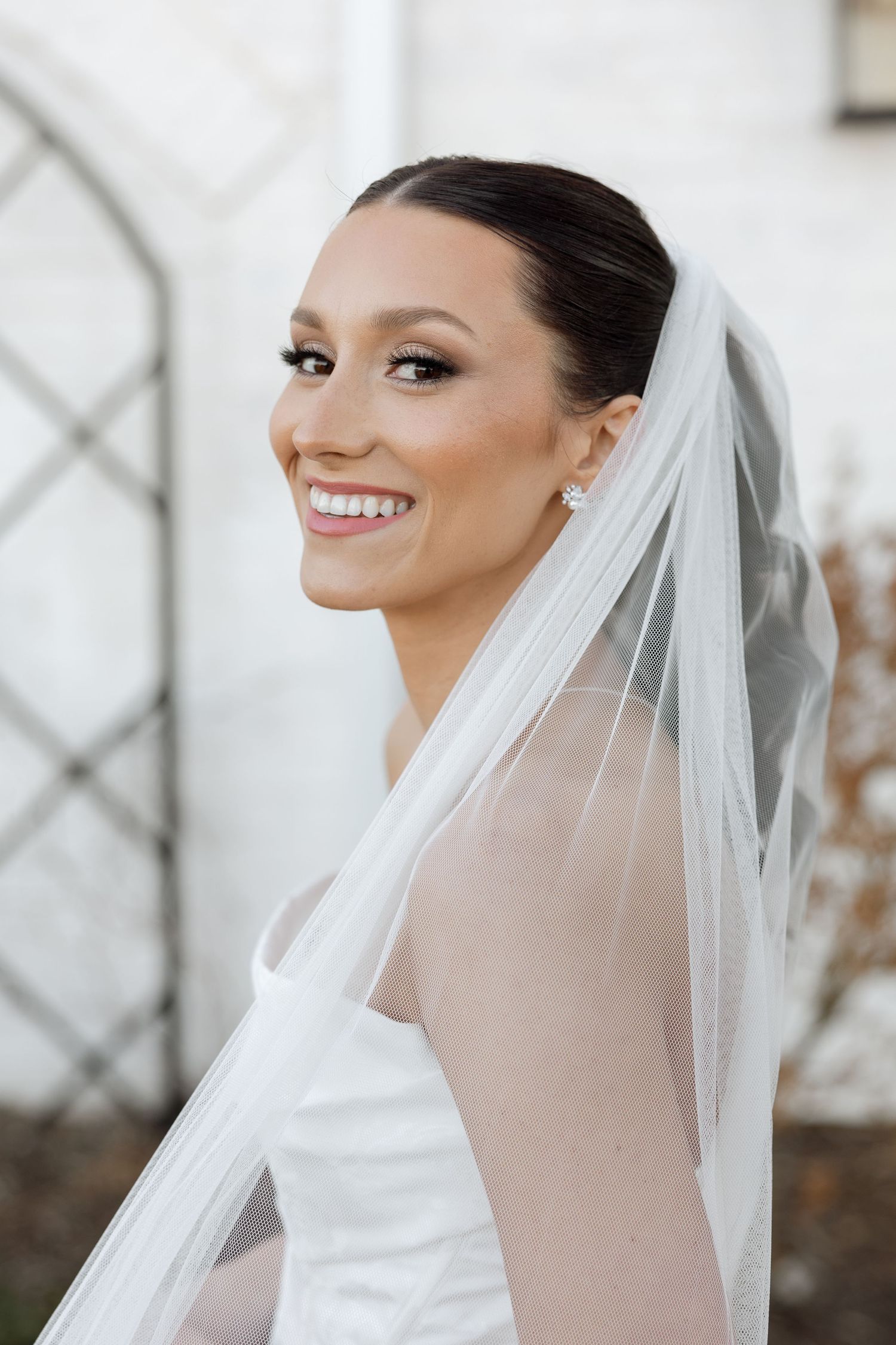 A radiant bride wearing a white wedding gown and flowing veil smiles brightly in an outdoor portrait.