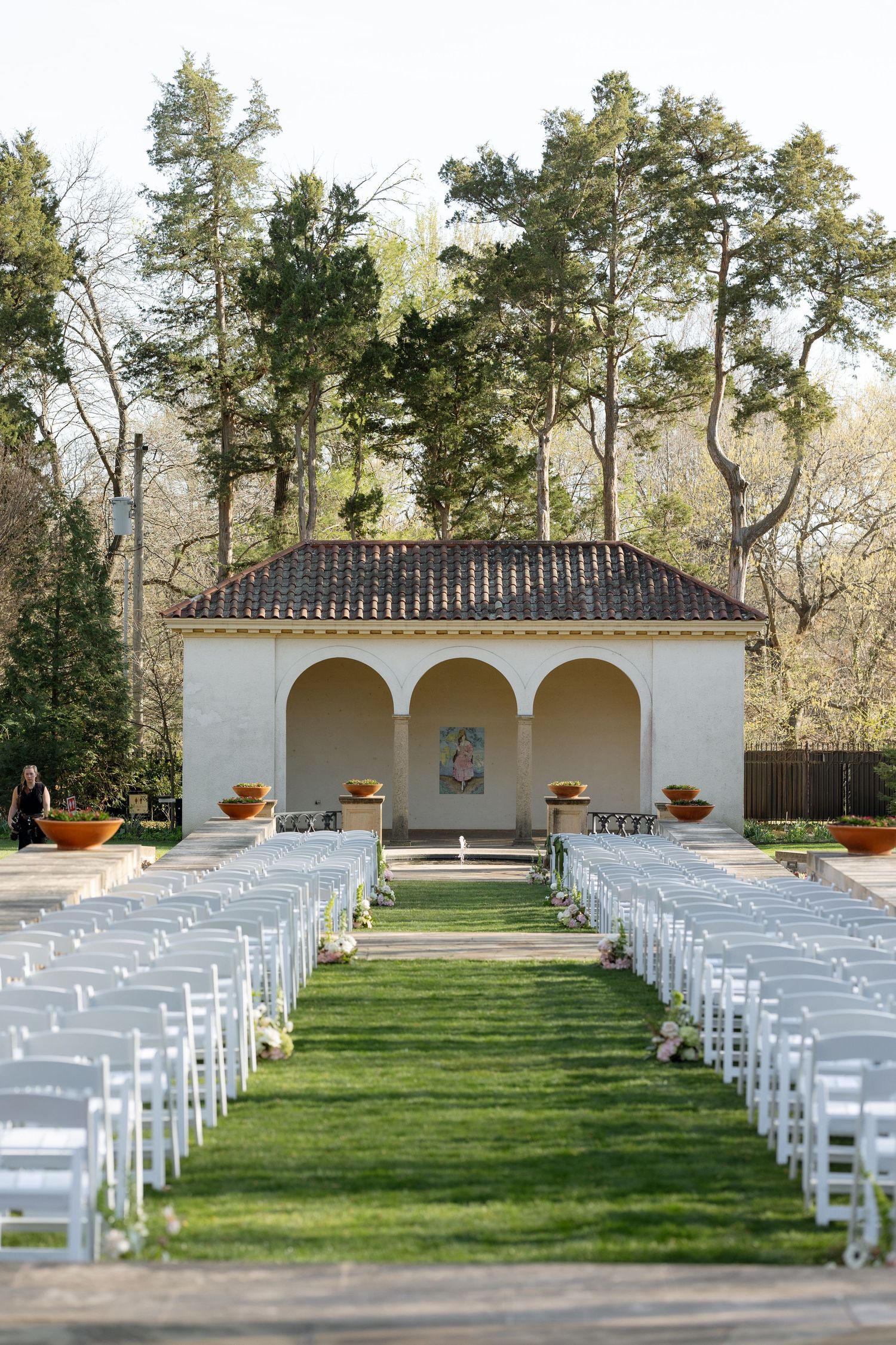 Outdoor wedding ceremony setup with white chairs on lawn facing arched pavilion surrounded by trees.