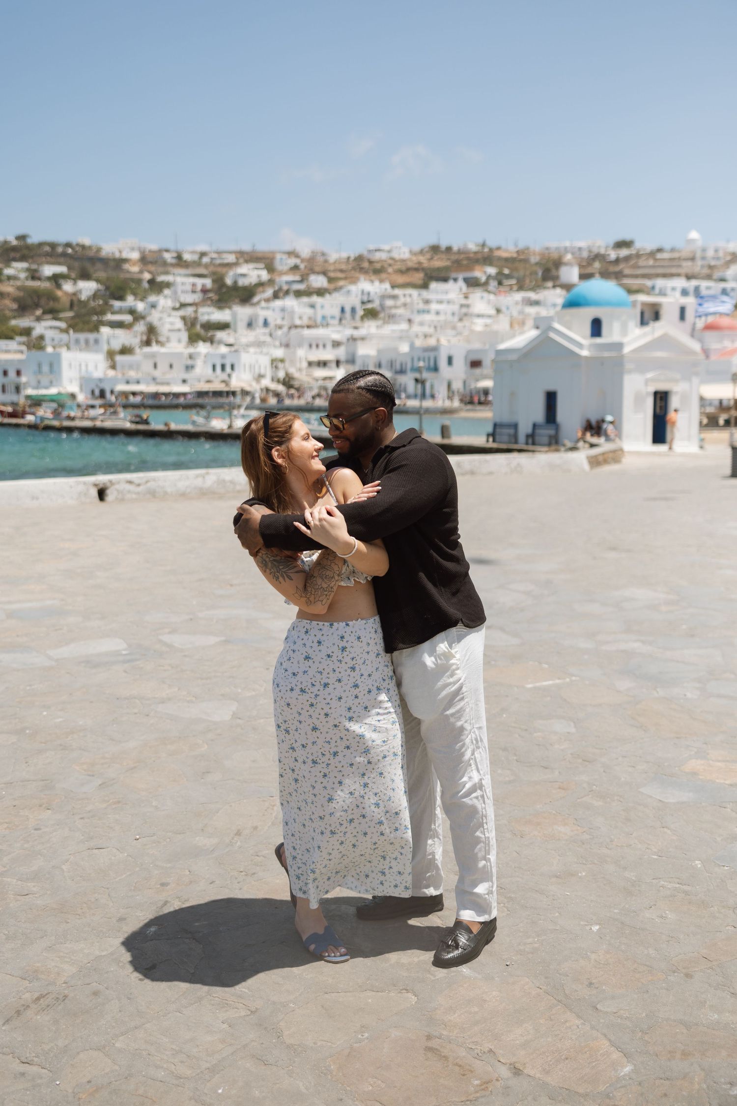 A couple dancing on a stone pier with traditional Greek architecture and blue domed church in background.