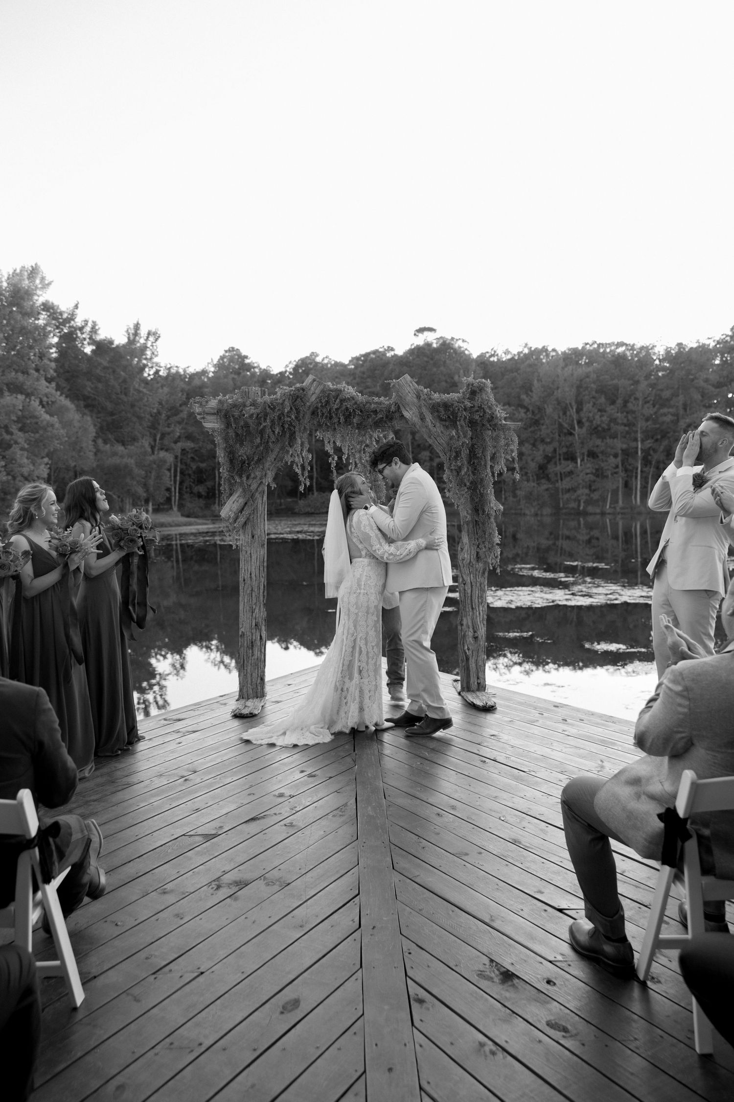 A black and white photo of a wedding ceremony on a wooden dock surrounded by guests.