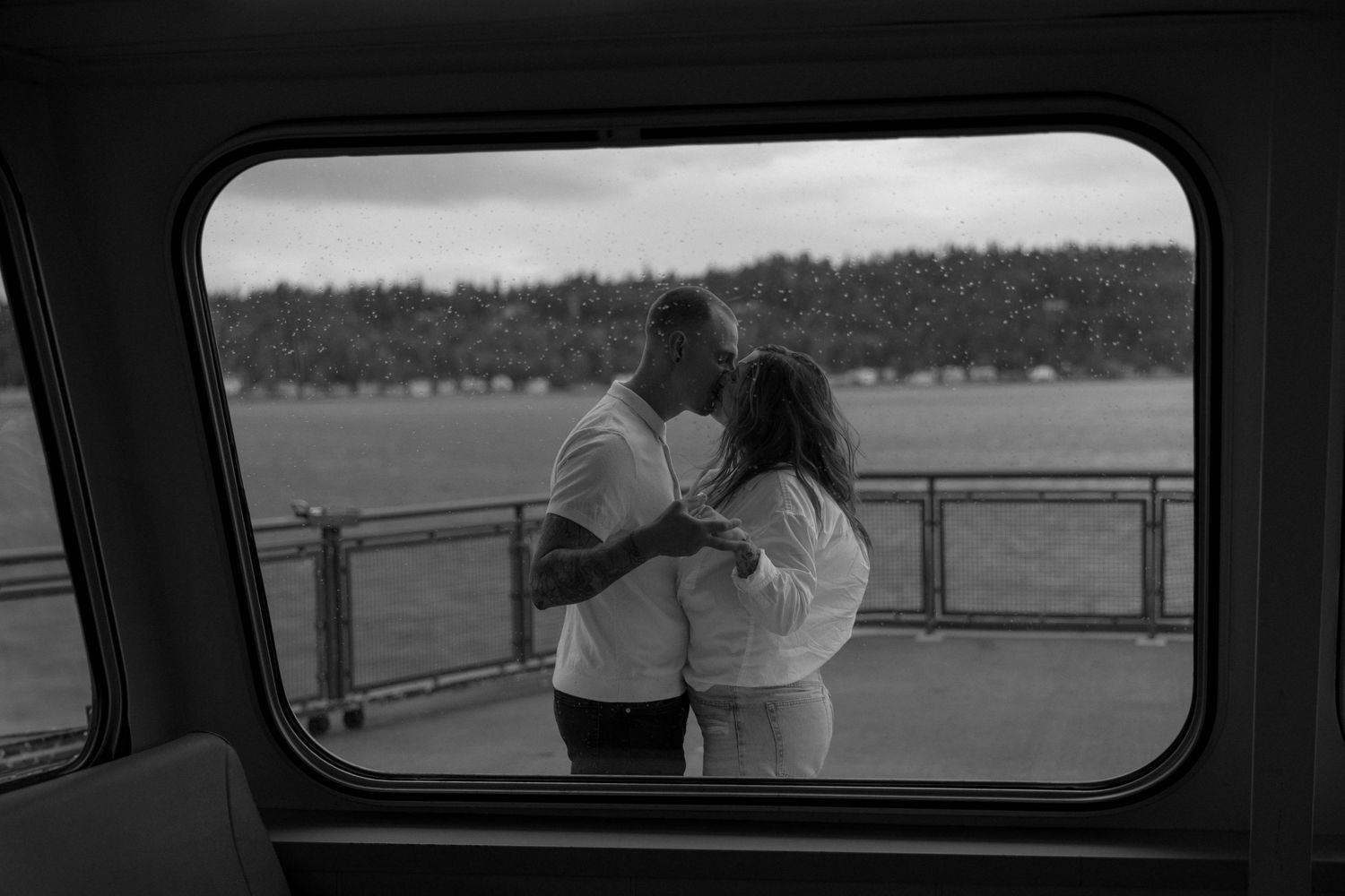 A romantic moment viewed through a ferry window as a couple embraces on the deck with water and trees in background.