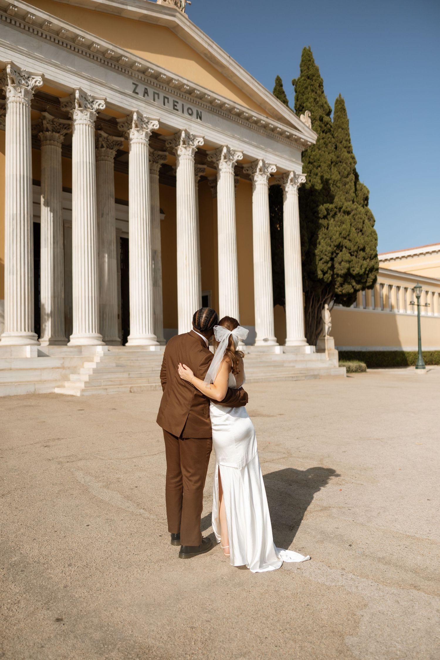 Wedding couple embraces in front of classical Zappeion building with grand columns.