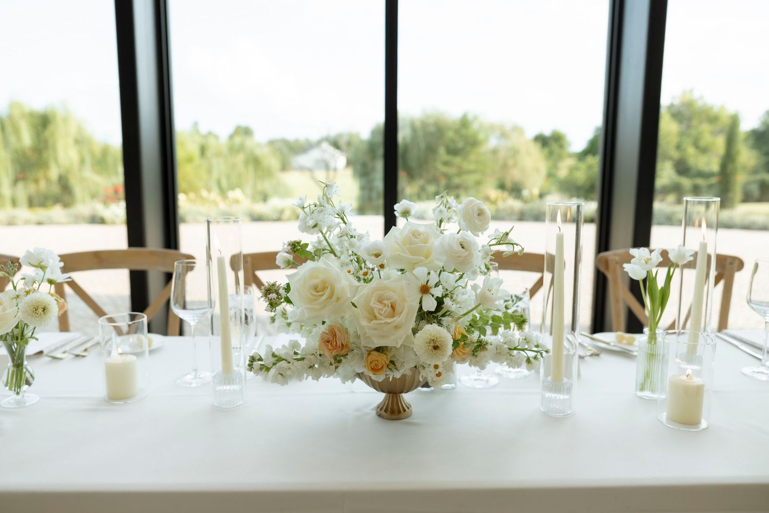 Long banquet tables with white floral centerpieces and cross-back chairs in a bright reception hall with panoramic windows.