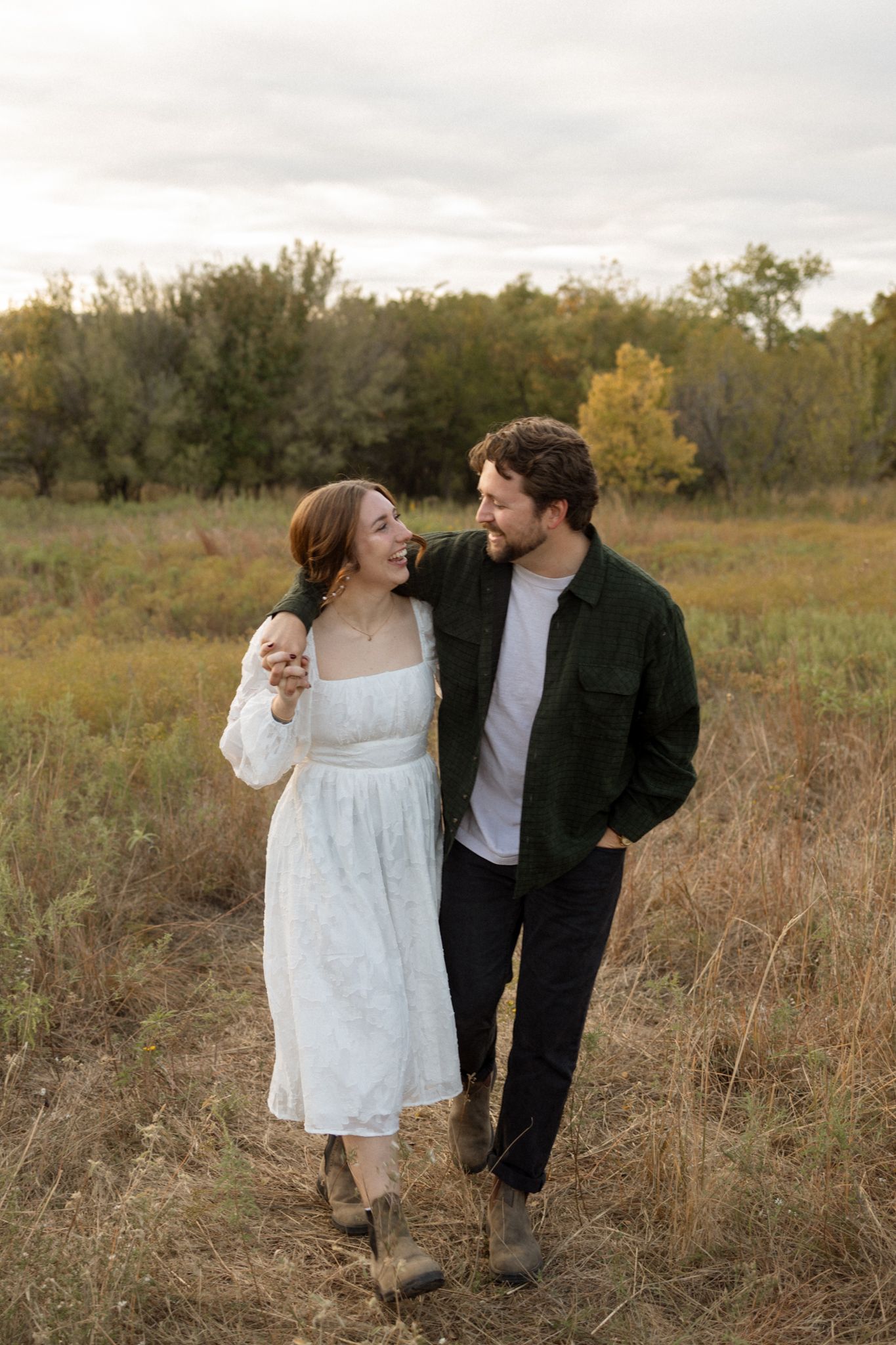 A couple walks together through a fall field, wearing a white dress and dark jacket.